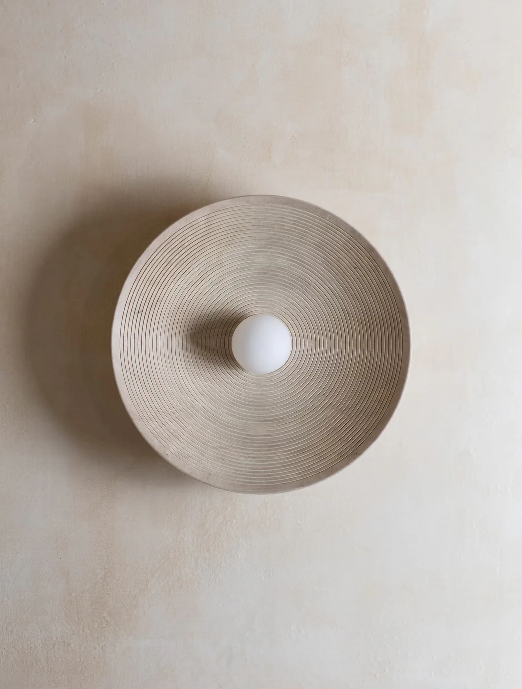 Minimalist circular pendant light fixture with concentric ribbed texture and central white sphere, suspended against neutral wall by Ryden Rizzo.