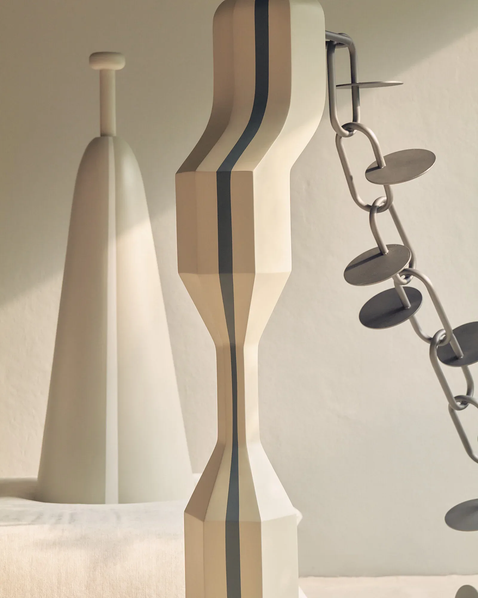 Modern sculptural ceramic and metal objects displayed against a neutral wall, featuring white vessels and striped forms with suspended metal disc elements, design by Saba Yazdjerdi.