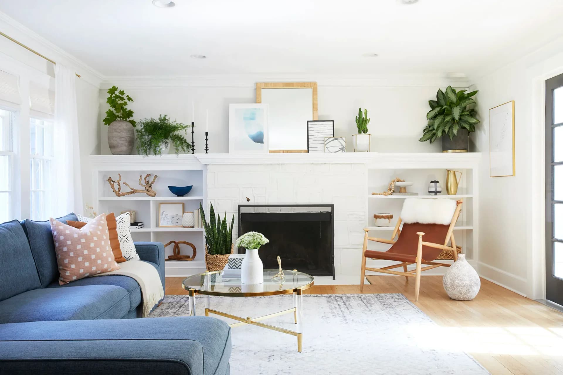 Bright living room with blue sofa, fireplace, floating shelves with plants and artwork, wooden chair, and light wood flooring by Sabrina Vroeg.
