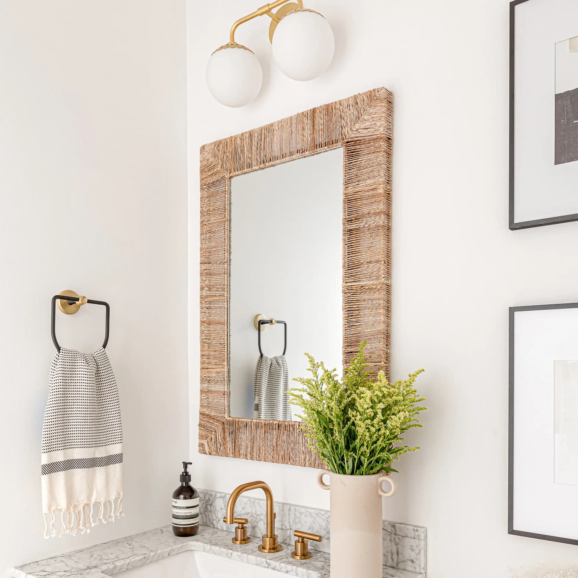 Modern bathroom with woven natural fiber mirror frame, marble countertop, brass faucet, and potted greenery by Sabrina Vroeg.