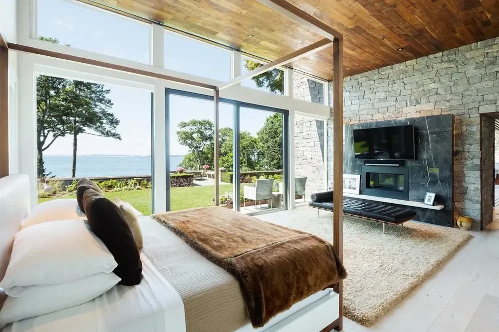 Modern bedroom with wooden canopy bed, white bedding, and expansive glass doors opening to waterfront views, stone fireplace accent wall, and ocean landscape by Salvador Juarez.