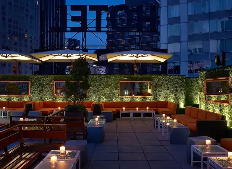 Evening rooftop terrace with orange seating, lush greenery, uplighting, and large umbrellas overlooking city skyline, designed by Salvador Juarez.