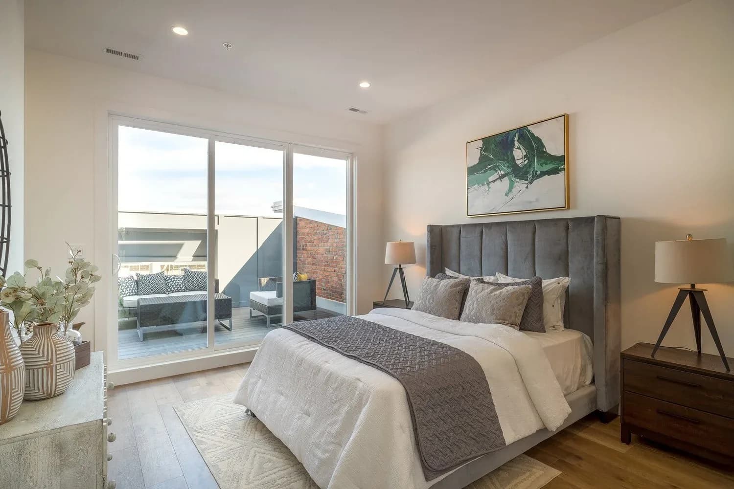 Modern bedroom with gray upholstered headboard, warm neutral palette, hardwood floors, and floor-to-ceiling windows overlooking a terrace by Sam Forman.