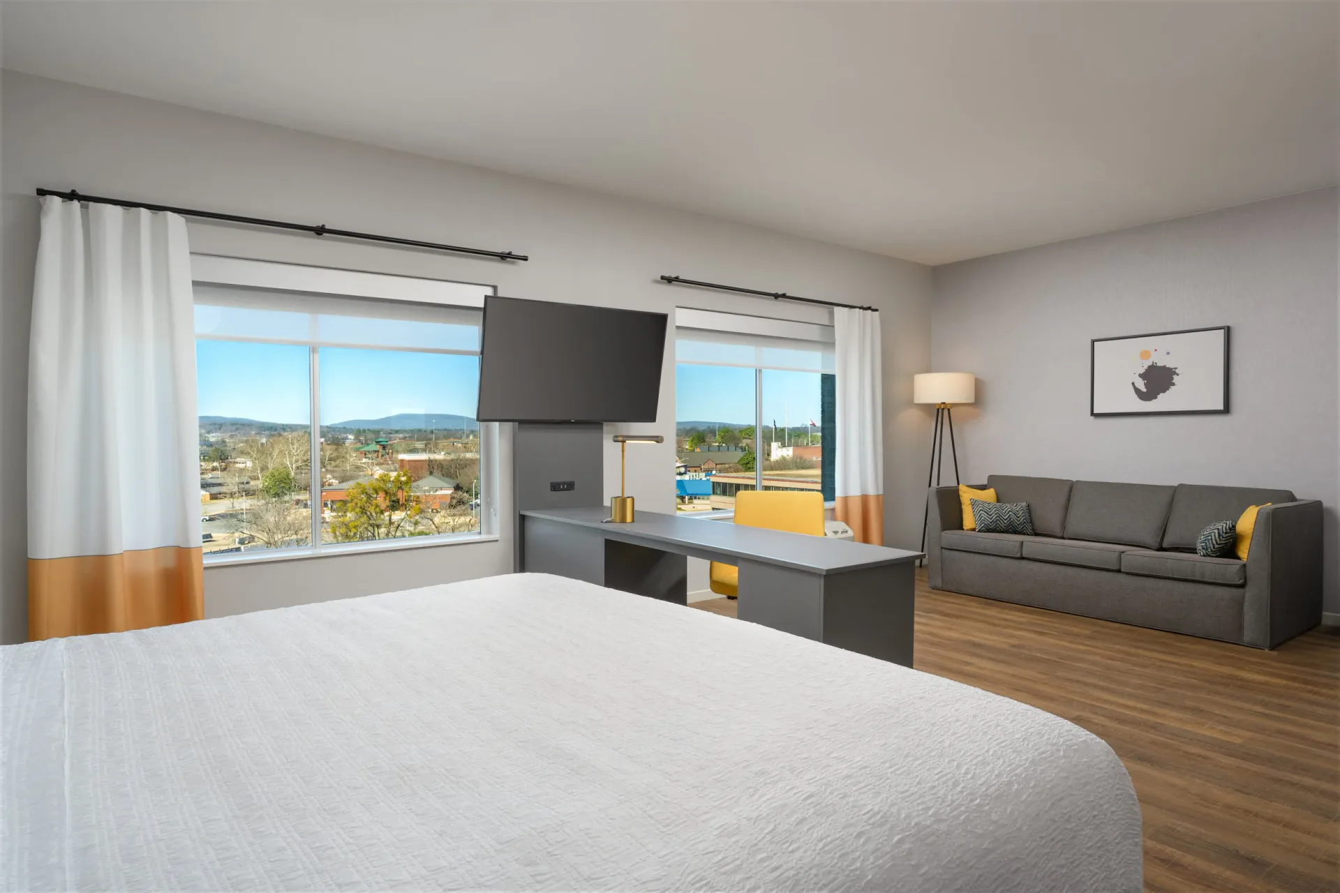 Modern hotel bedroom with gray sofa, mounted TV, panoramic city views, and wood flooring by Sam Forman.