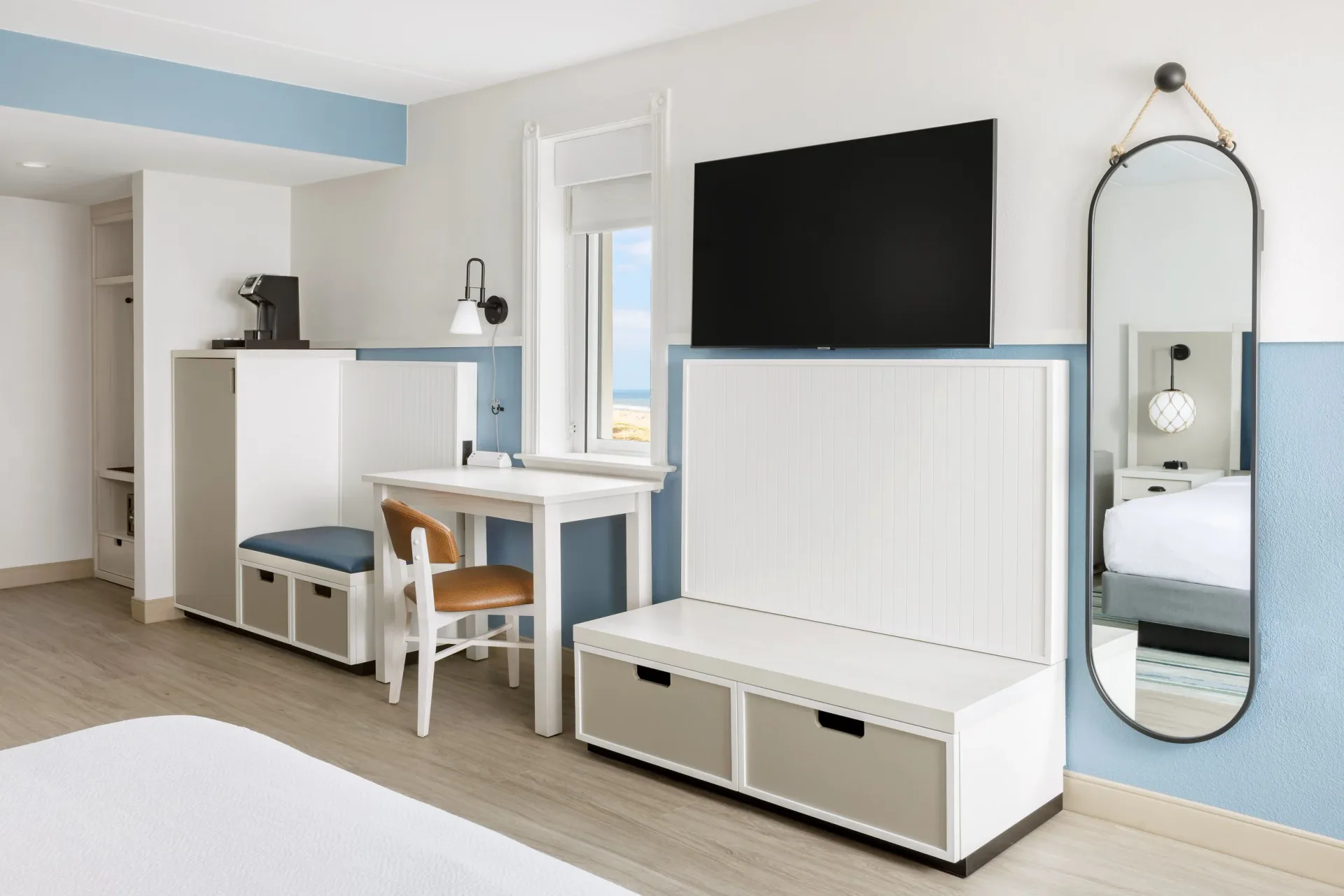 Modern hotel or residential bedroom with white and blue two-tone walls, integrated TV unit with storage, desk area, and round mirror by Sam Forman.