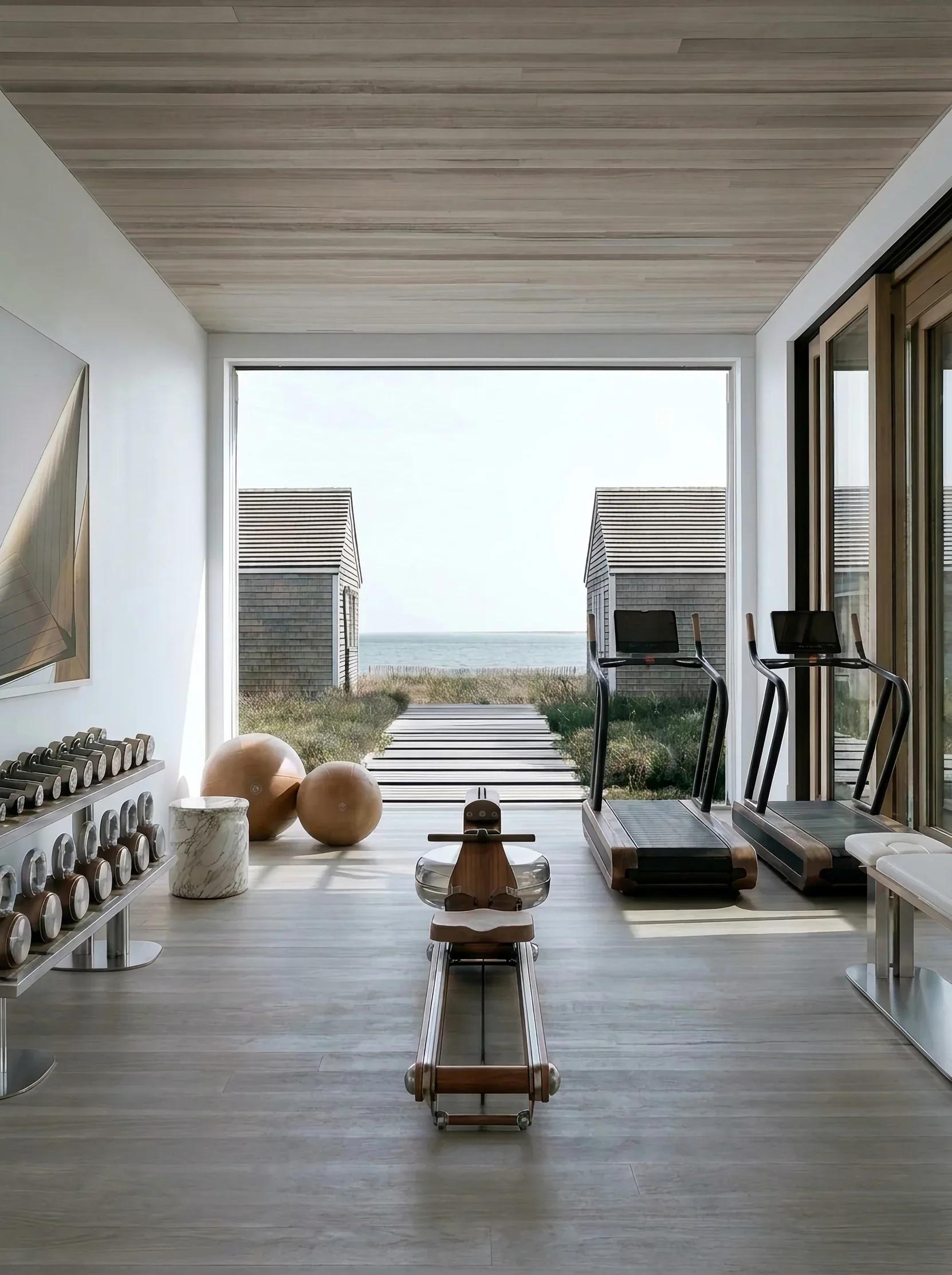 Minimalist coastal spa interior with polished concrete floors, wood ceiling, and floor-to-ceiling windows overlooking beach, featuring sculptural wood and ceramic objects by Sam Kimball.
