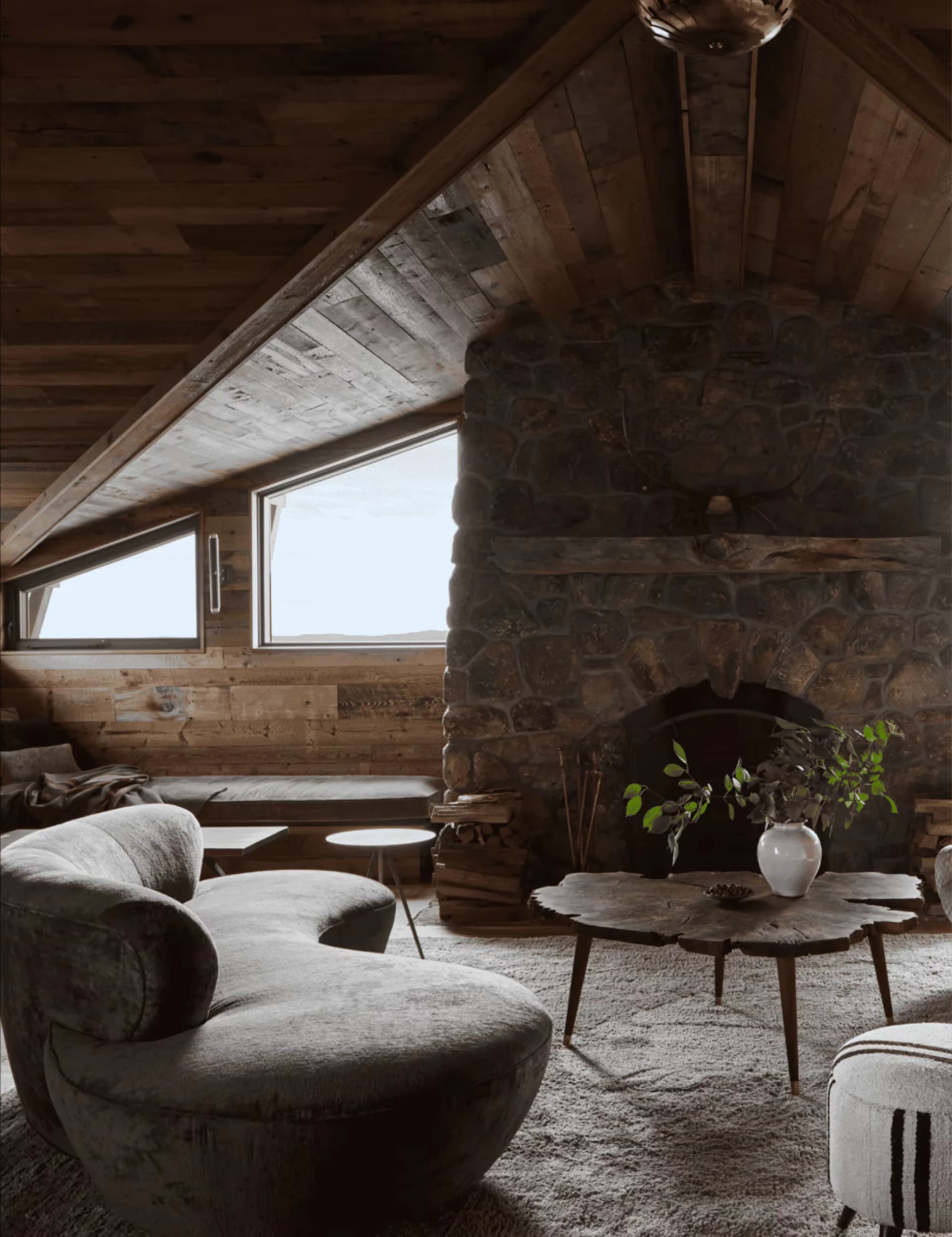 Rustic alpine cabin interior with vaulted wooden ceiling, stone fireplace, curved upholstered seating, and panoramic windows overlooking mountains by Samantha Hemsworth.