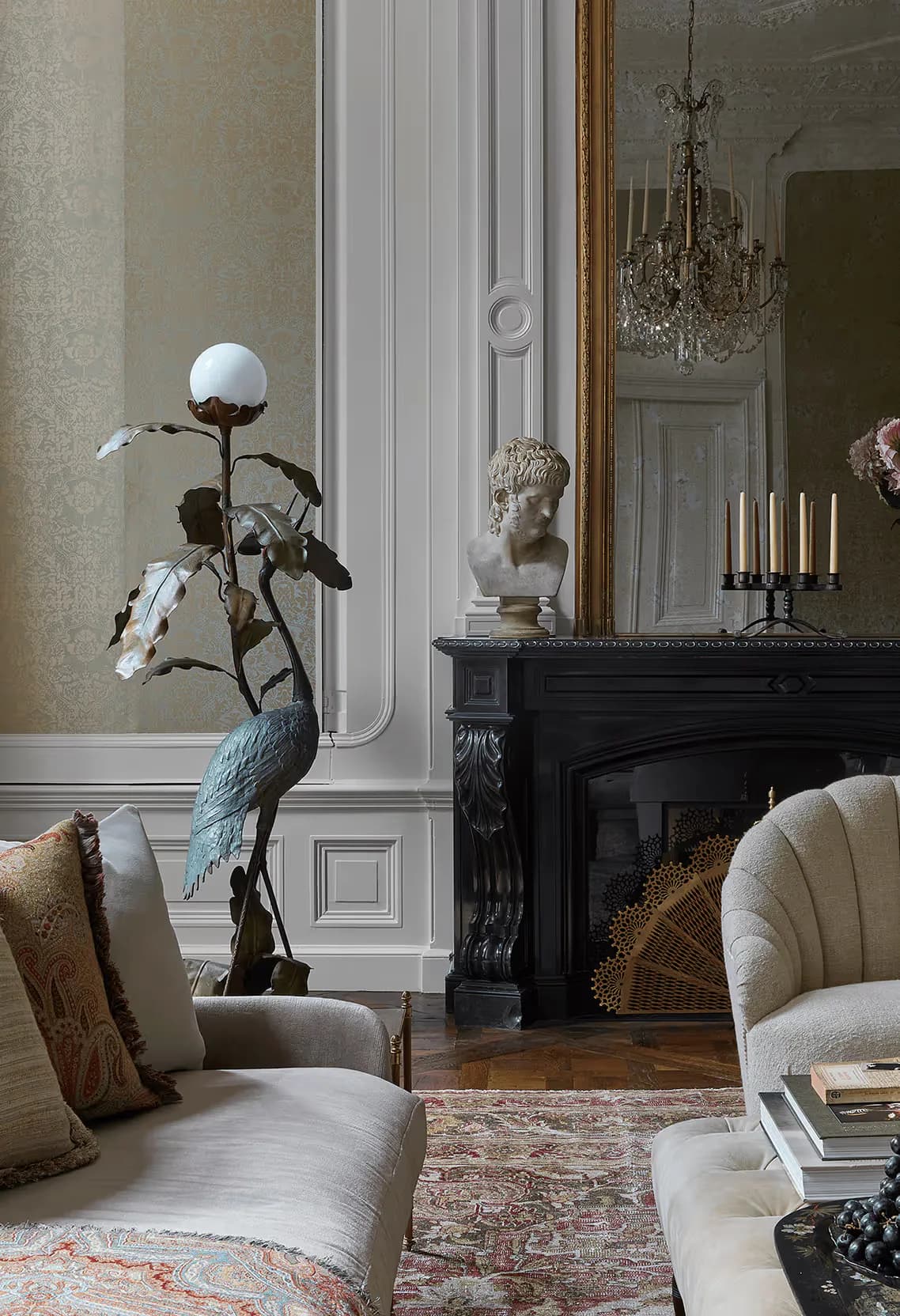 Elegant living room with cream curved seating, black marble fireplace, ornate mirror, crystal chandelier, and sculptural blue bird figurine by Samantha Hemsworth.