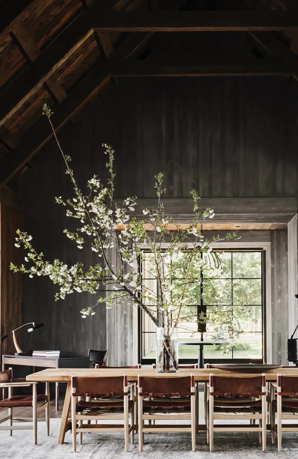 Modern dining space with dark walls, wooden beams, natural light, tall floral arrangement, and mid-century chairs by Samantha Hemsworth.