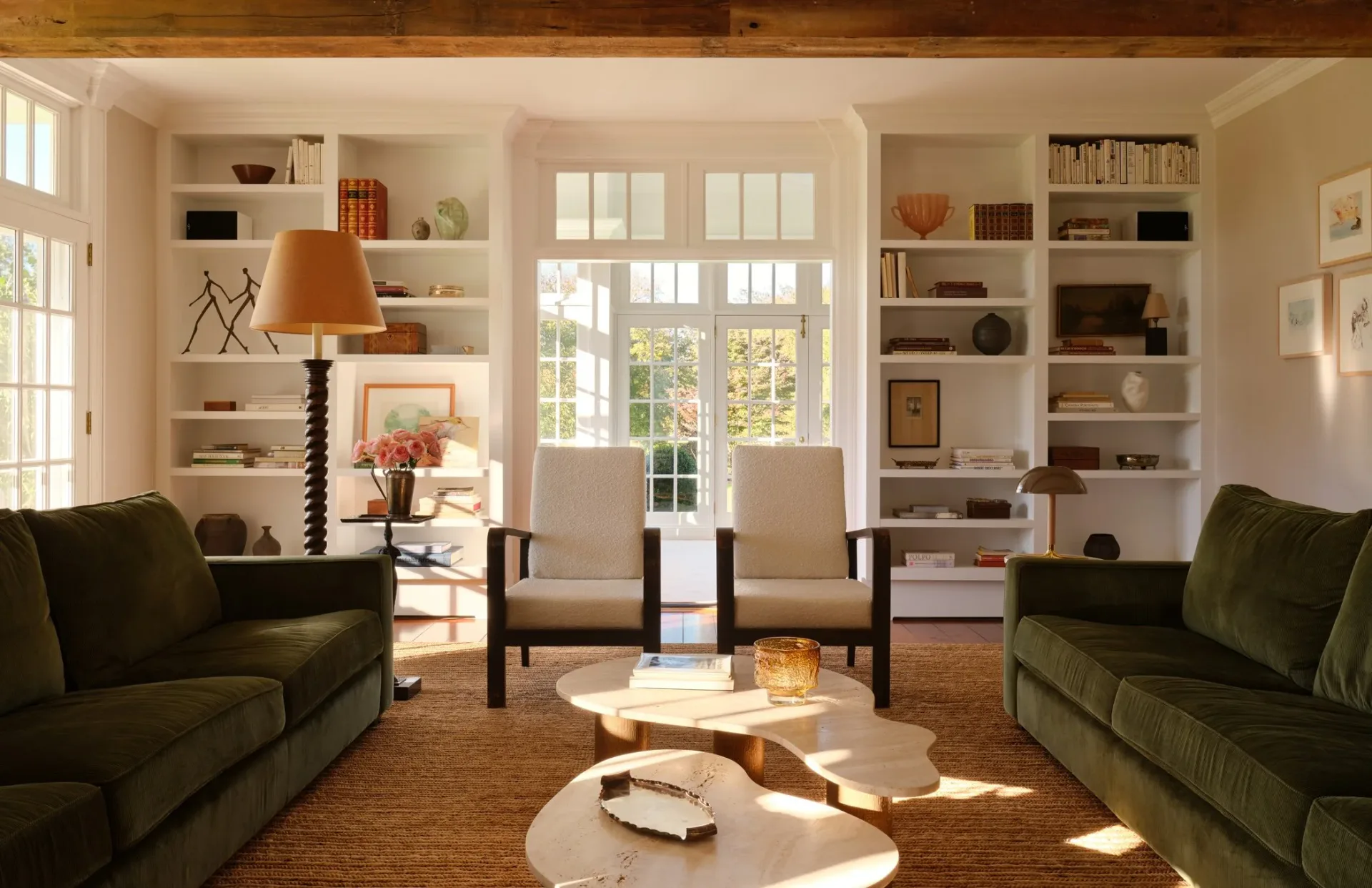 Bright living room with cream built-in shelving, two green upholstered sofas, neutral seating chairs, light wood flooring, and french doors opening to garden, designed by Samantha Tannehill.