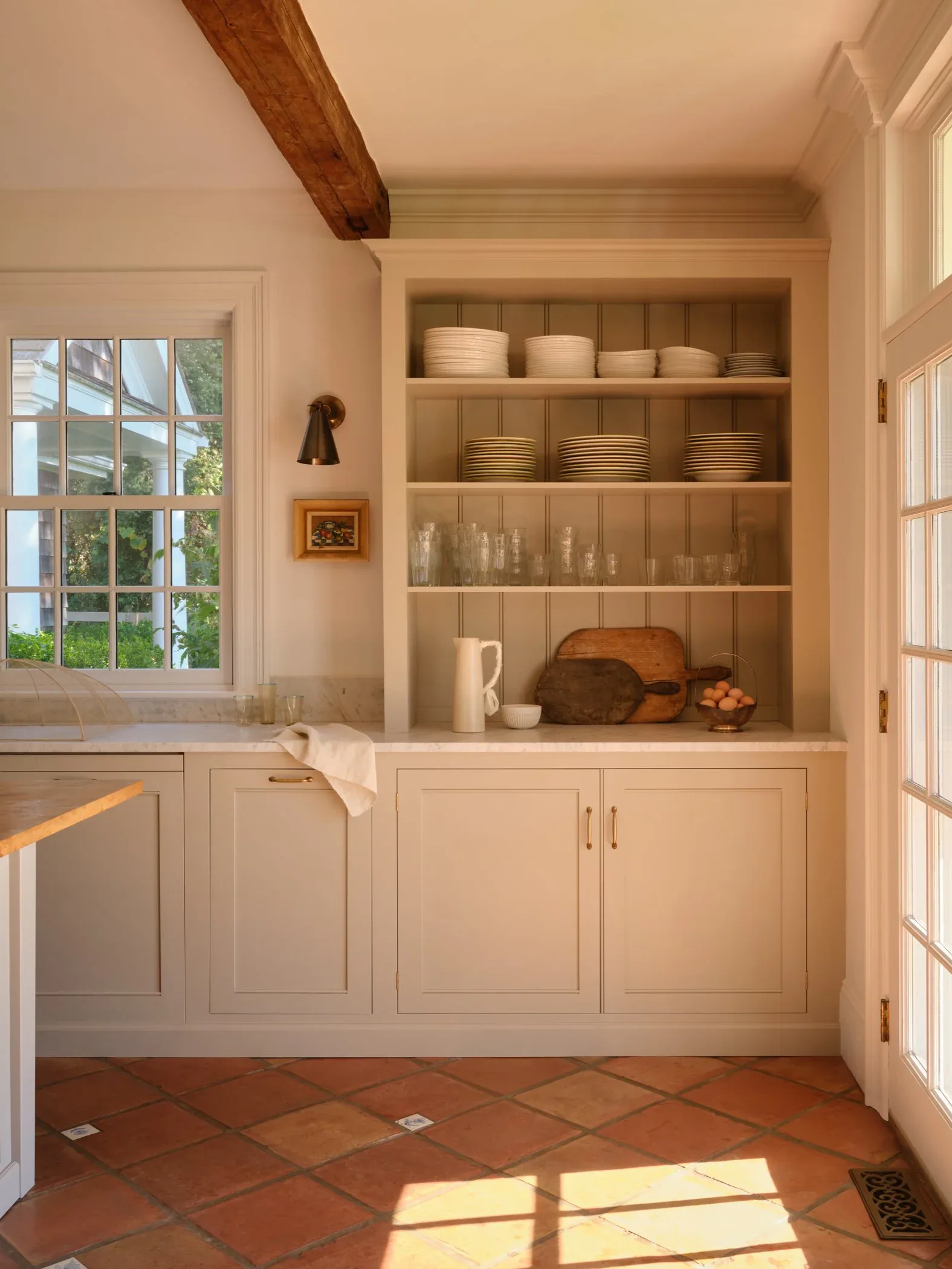 Cream-painted kitchen with open shelving displaying dinnerware, terracotta tile flooring, exposed wood beam, and natural light from multi-paned windows by Samantha Tannehill.