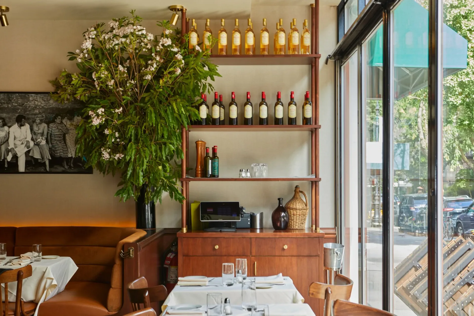 Warm-toned restaurant dining room with wooden shelving, wine bottles, leather seating, and large windows overlooking a street by Samantha Tannehill.