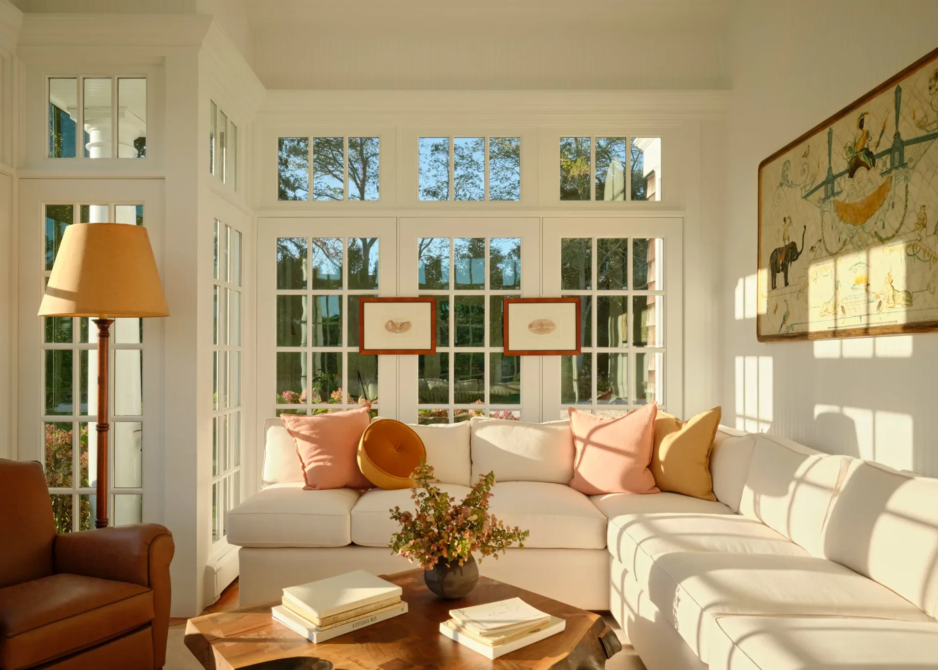 Sunlit living room with cream upholstered seating, coral accent pillows, extensive windows, wood furnishings, and decorative Asian artwork by Samantha Tannehill.
