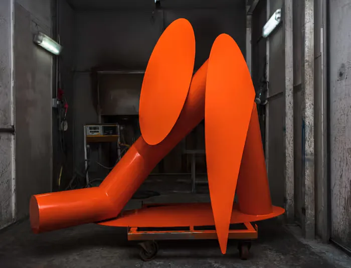 Bold orange sculptural installation with curved geometric forms mounted on a wheeled platform in an industrial gallery space by Samuel Ross.