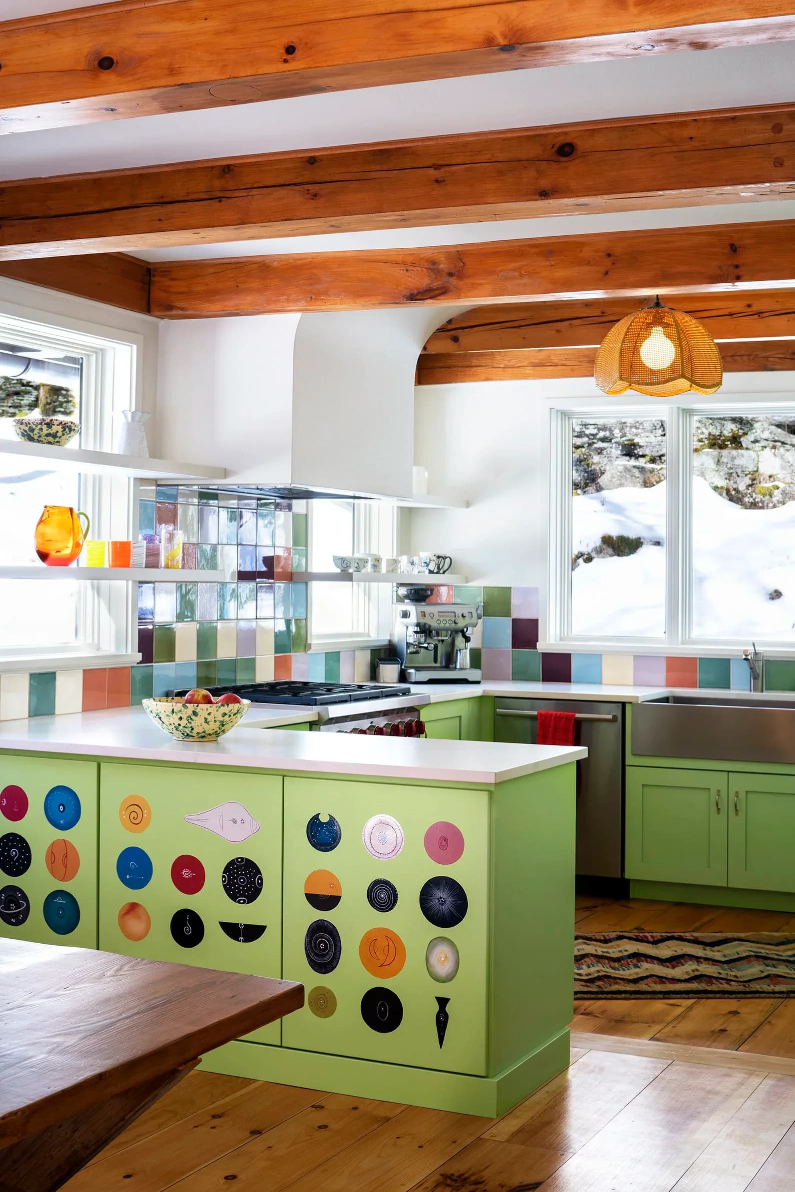 Bright kitchen with lime-green cabinetry featuring colorful decorative plates, wooden beam ceilings, multicolored tile backsplash, and large windows designed by Sara Bengur.