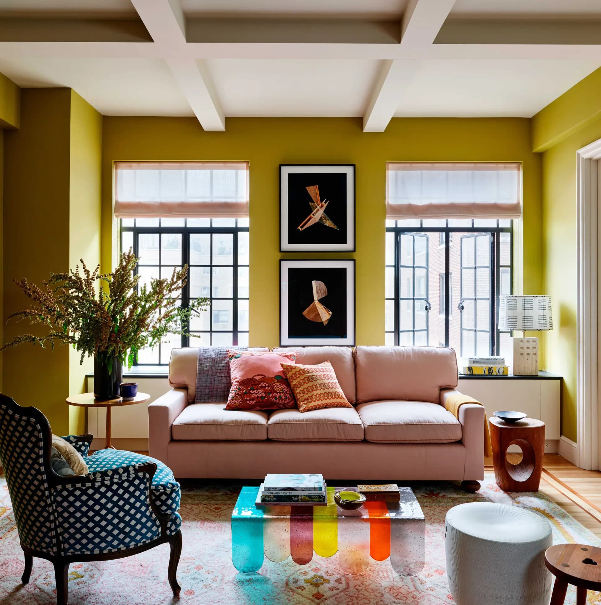 Vibrant yellow living room with mauve sofa, colorful striped ottoman, black patterned armchair, and framed artwork, designed by Sara Bengur.