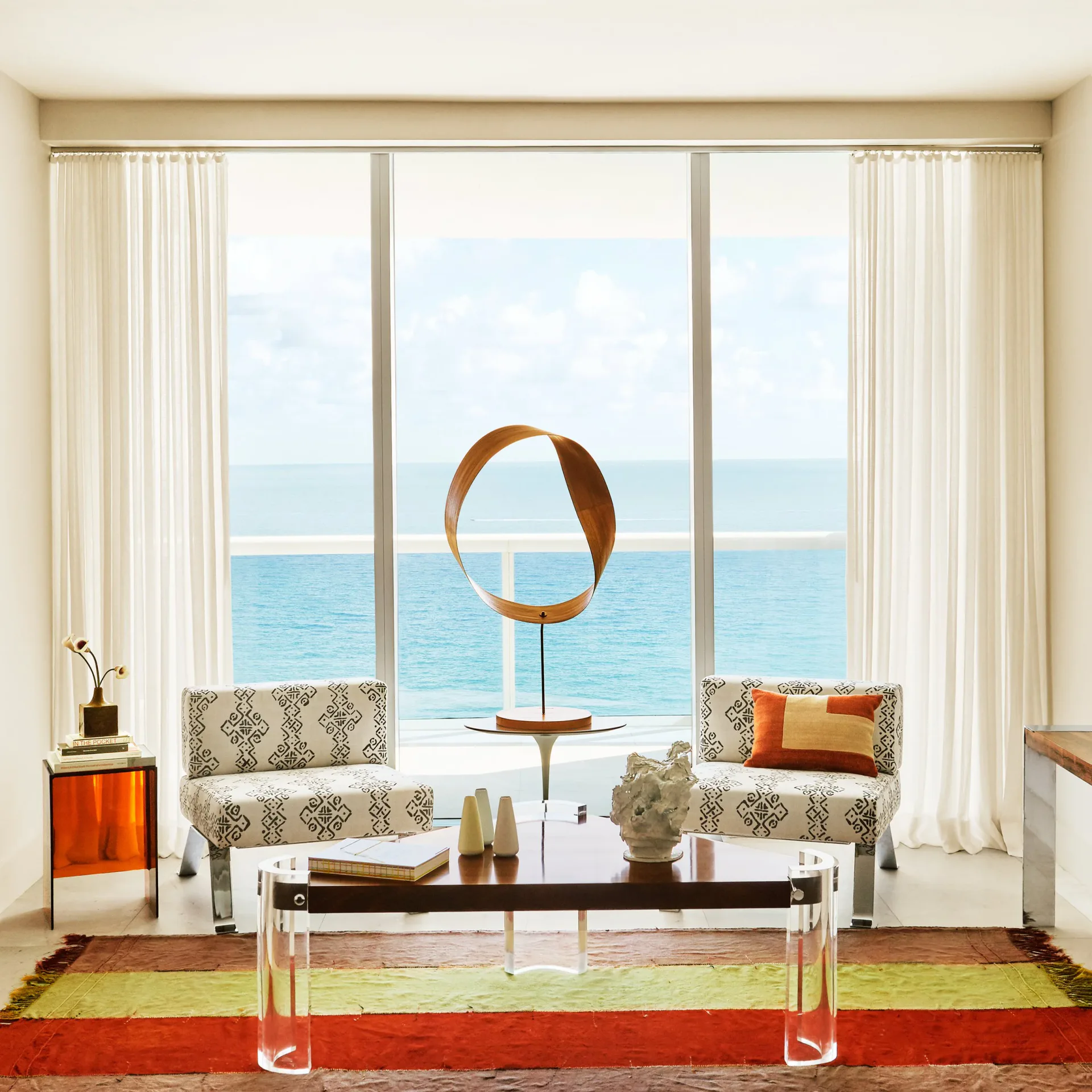 Coastal living room with floor-to-ceiling windows framing ocean views, neutral furnishings, patterned seating, sculptural bronze accent piece, and red area rug by Sara Bengur.