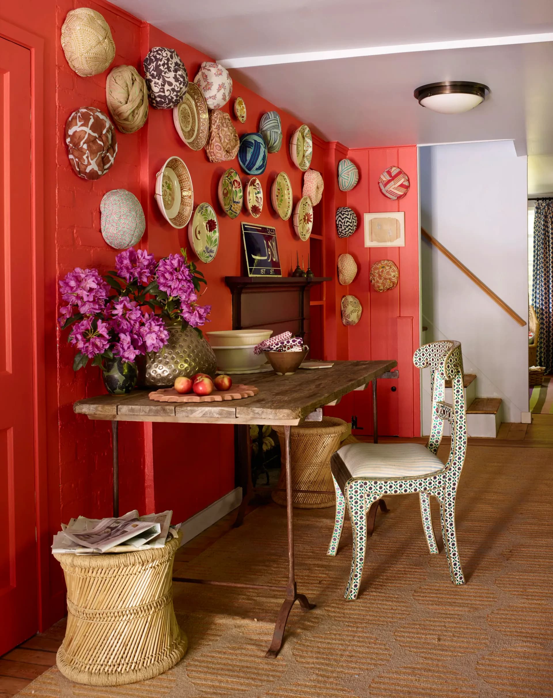 A vibrant red entryway with woven plate wall art, wooden console table, magenta flowers, and patterned chair by Sara Bengur.