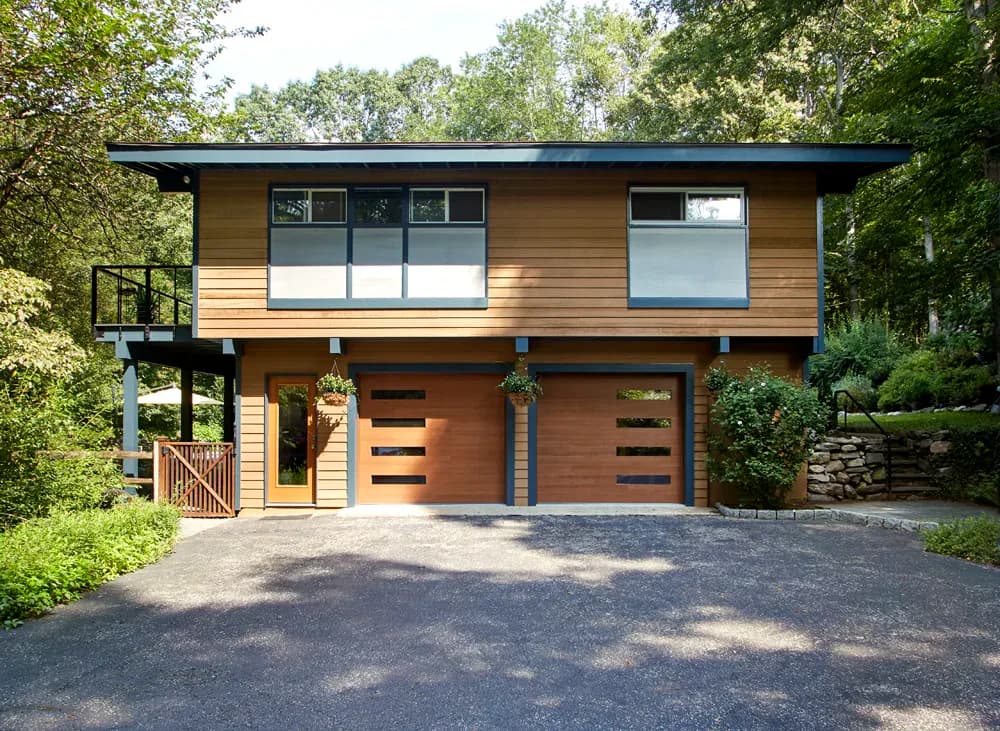 Modern two-story residential home with warm wood siding, large glazed windows, and wood-paneled garage doors set among mature trees by Sara Jazayeri.
