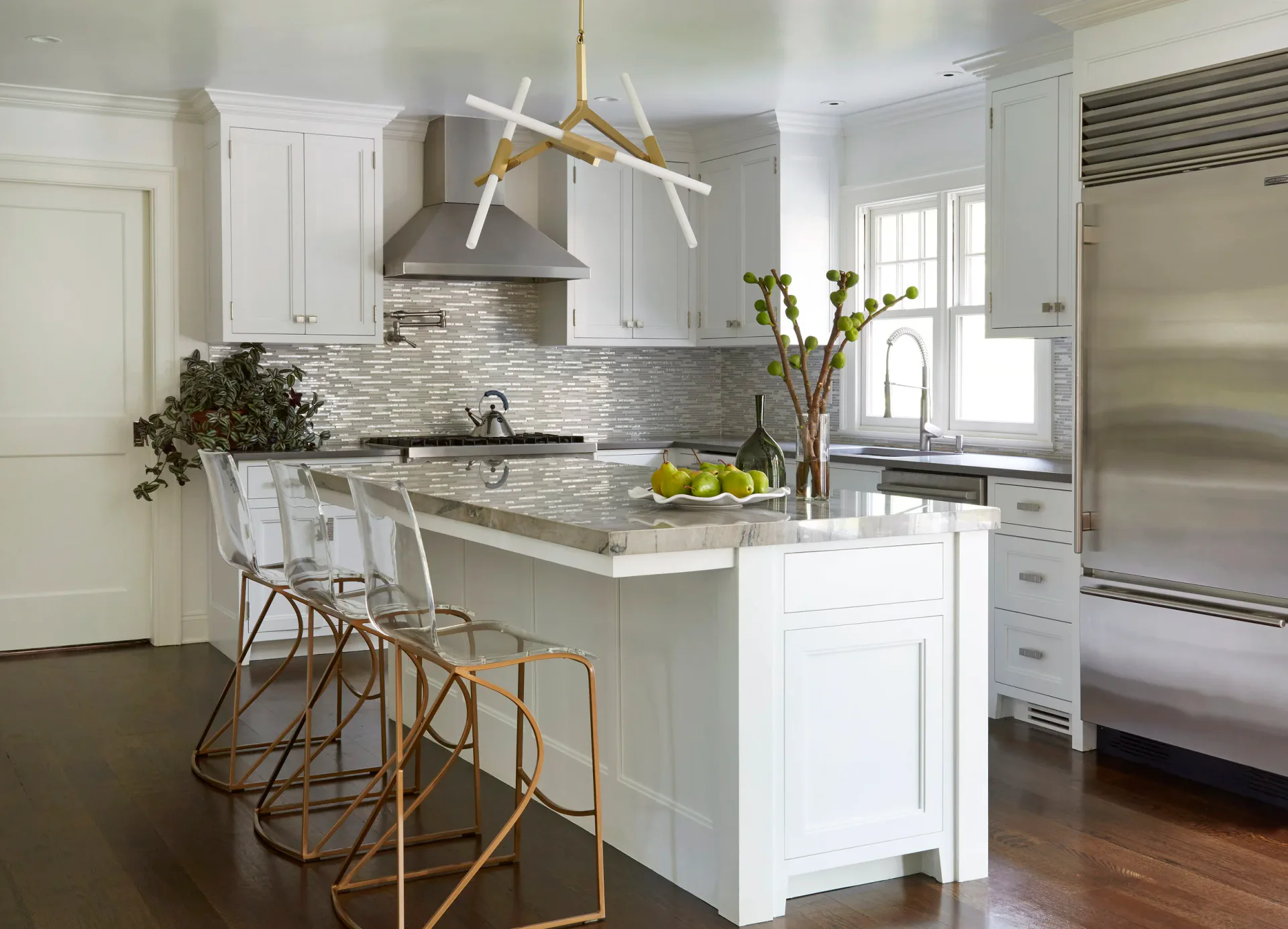Modern white kitchen with gray marble countertops, brass pendant lighting, woven bar stools, and stainless steel appliances by Sara Jazayeri.