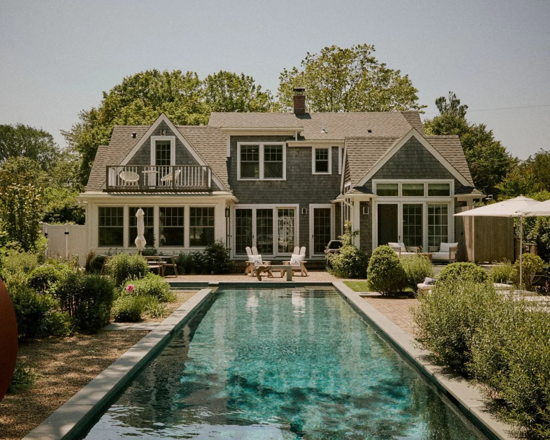 Modern farmhouse exterior with dark-framed windows, gabled roofline, and linear swimming pool surrounded by landscaping by Sarah Witkin.