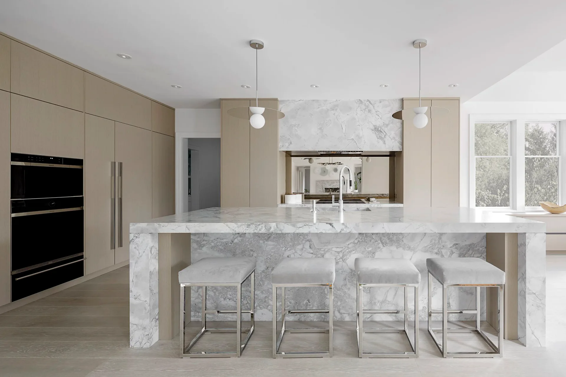 Modern kitchen with marble waterfall island, light wood cabinetry, and four upholstered bar stools by Sarah Witkin.