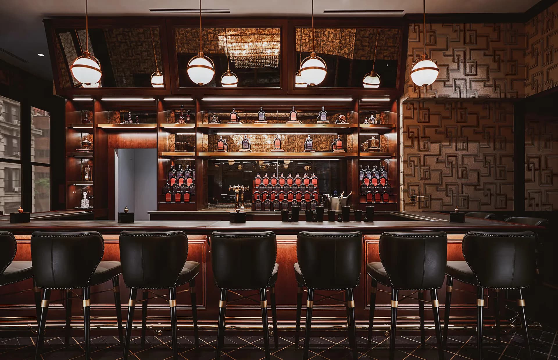 Upscale bar interior with dark wood cabinetry, brass pendant lights, black leather bar stools, and warm brick-textured walls designed by Sarahg Gorski.