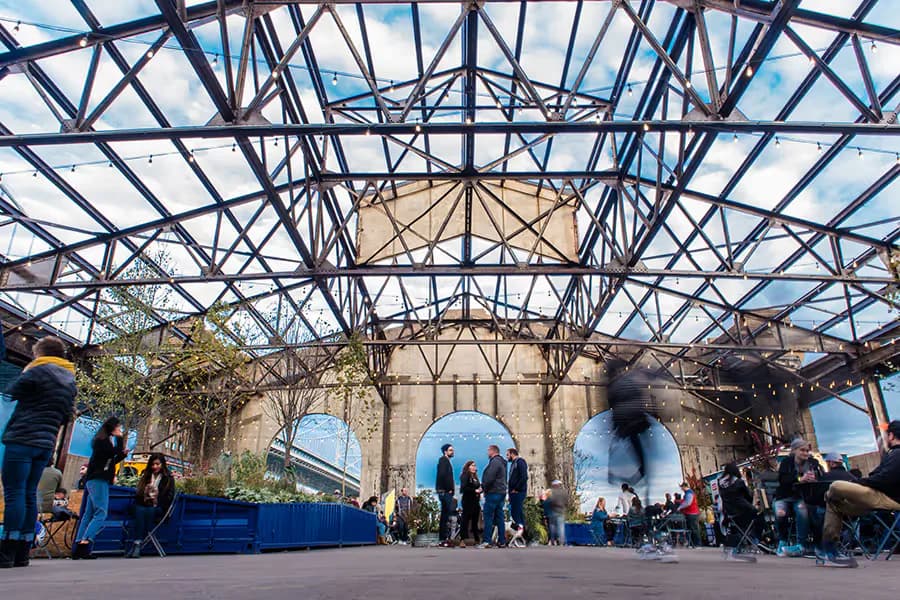 A modern steel-framed pavilion with glass roof panels shelters a historic stone building, creating an open-air gathering space with industrial geometry and natural light by Sarahg Gorski.