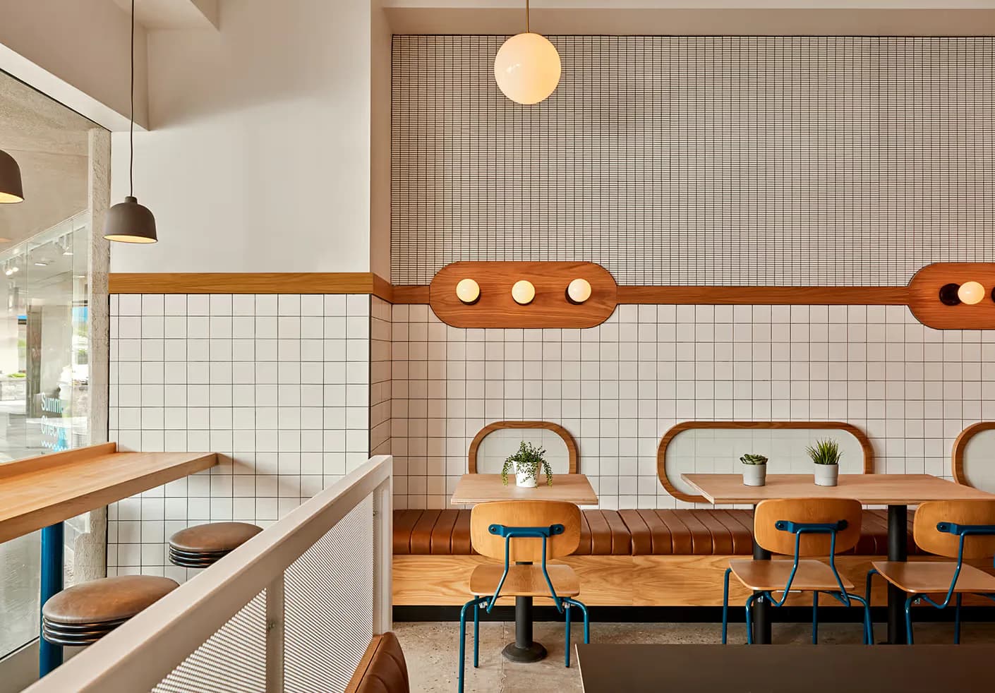 Warm-toned café interior with white subway tile walls, wooden banquette seating, metal chairs, and curved wood architectural details by Sarahg Gorski.