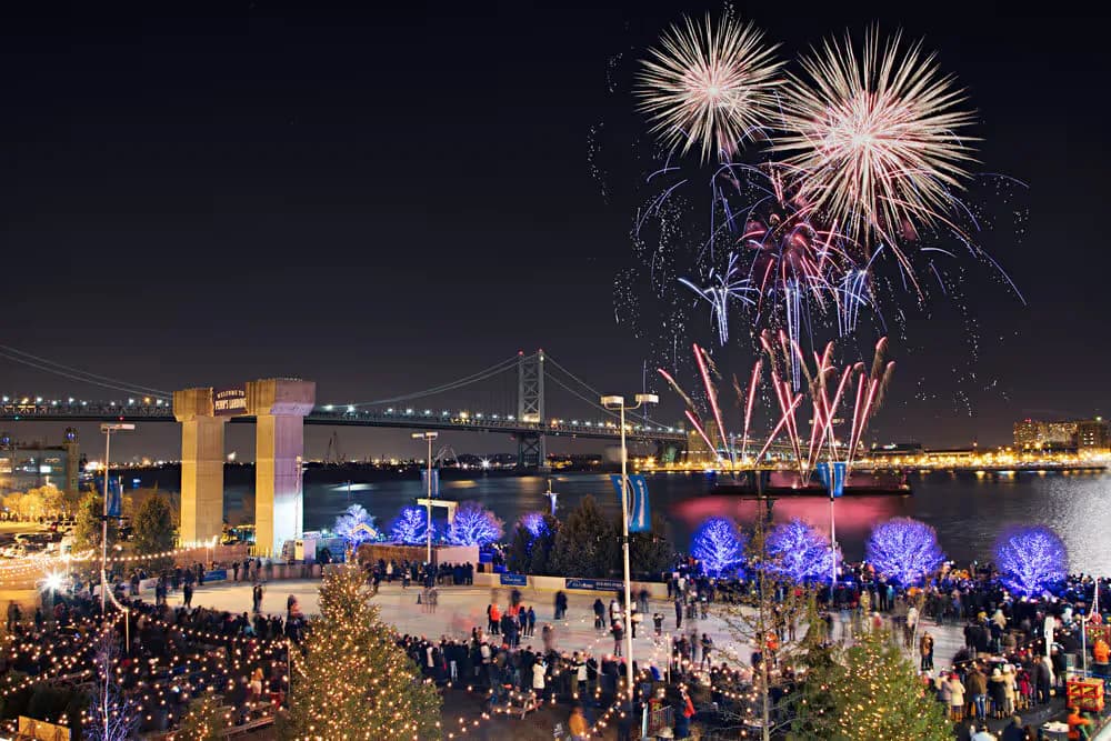 Waterfront plaza at night features illuminated architectural structures, blue-lit trees, crowds of visitors, and fireworks exploding above a bridge, designed by Sarahg Gorski.