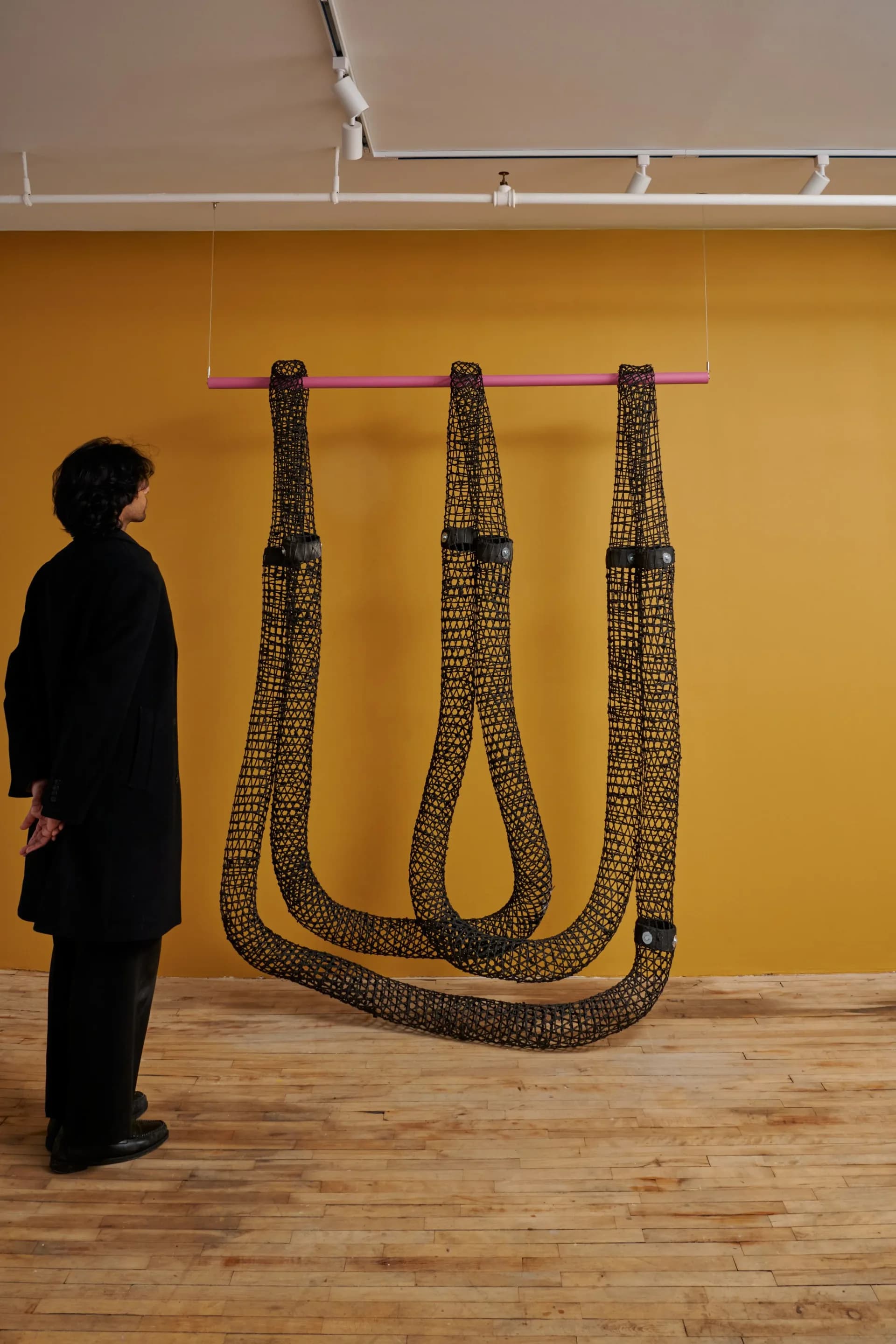 Woven rope installation suspended from pink rod against mustard yellow wall, with viewer in black coat observing the sculptural piece by Sarita Westrup.