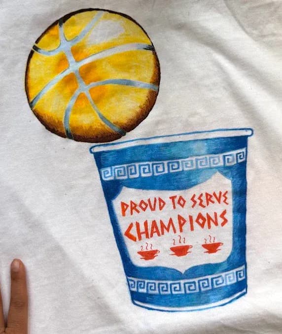 Hand-painted design on white fabric featuring a golden basketball with blue lines and a blue cup labeled 'Proud to Serve Champions' with decorative Greek pattern borders by Sasha Arijanto.