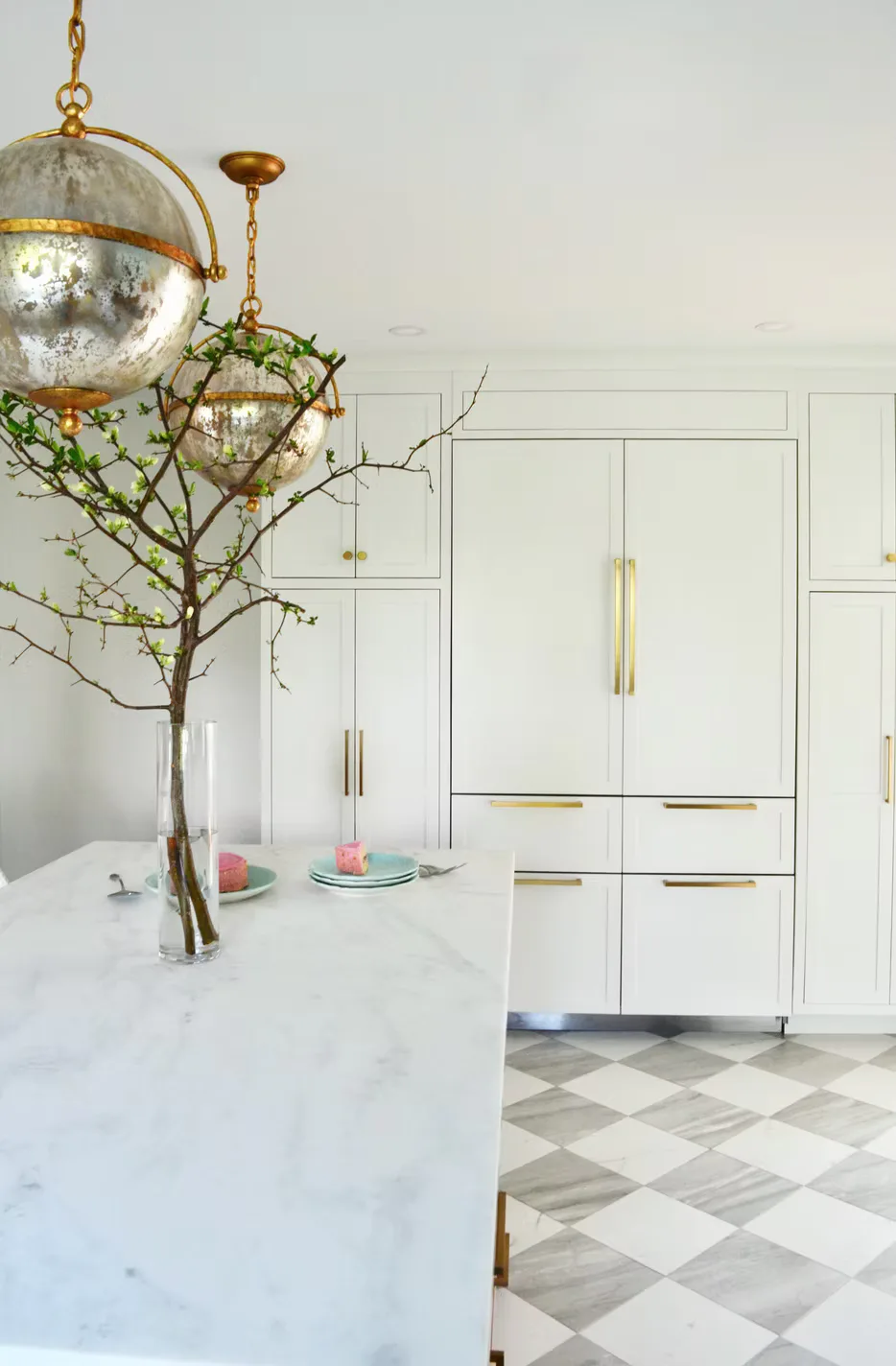 Modern white kitchen with marble countertop, brass globe pendant light, and white cabinetry with brass hardware, featuring a geometric black and white tile floor by Schuyler Johnson.