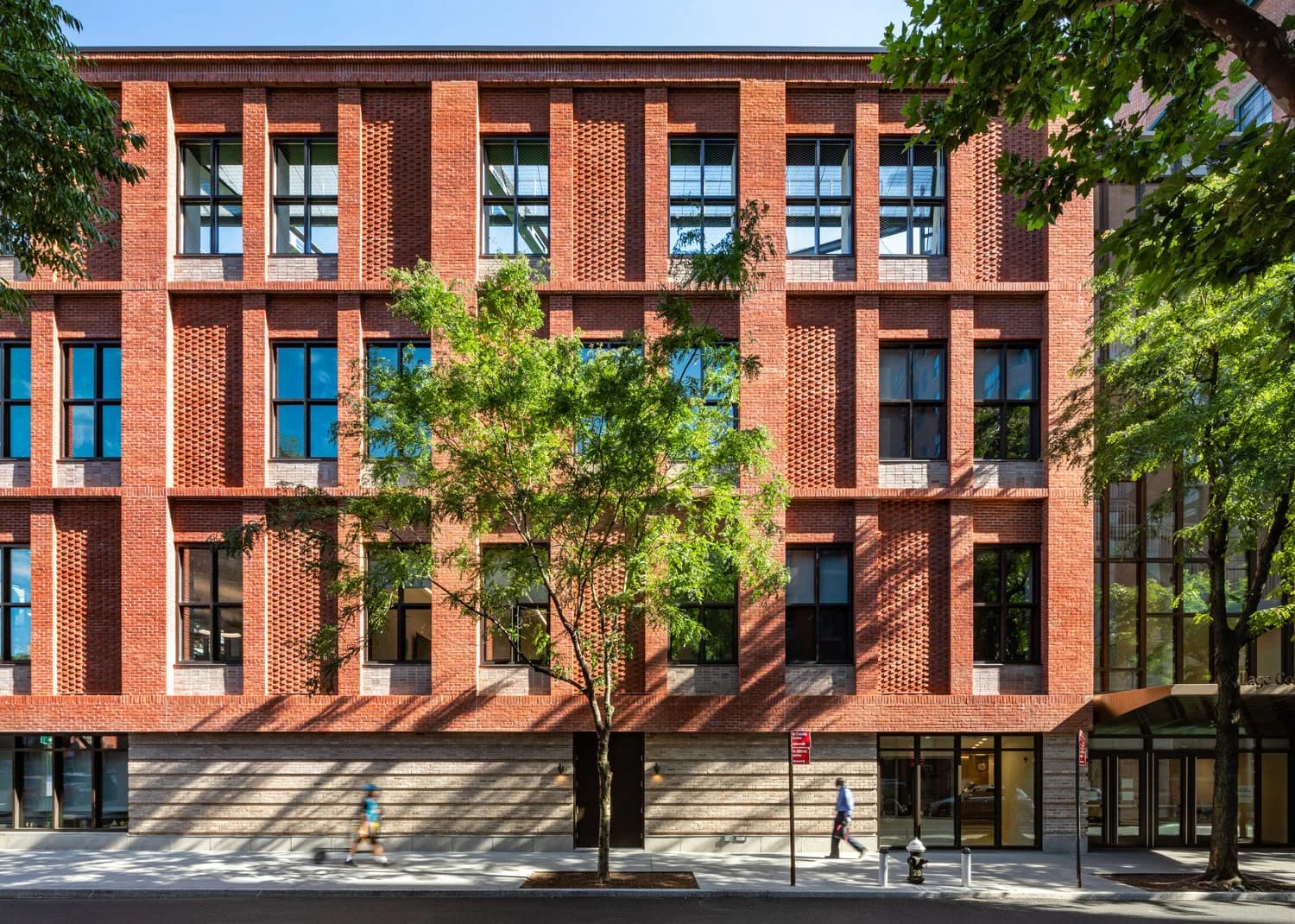 Red brick contemporary residential building with vertical ribbed detailing, blue-framed windows, and integrated landscaping by Scott Demel.