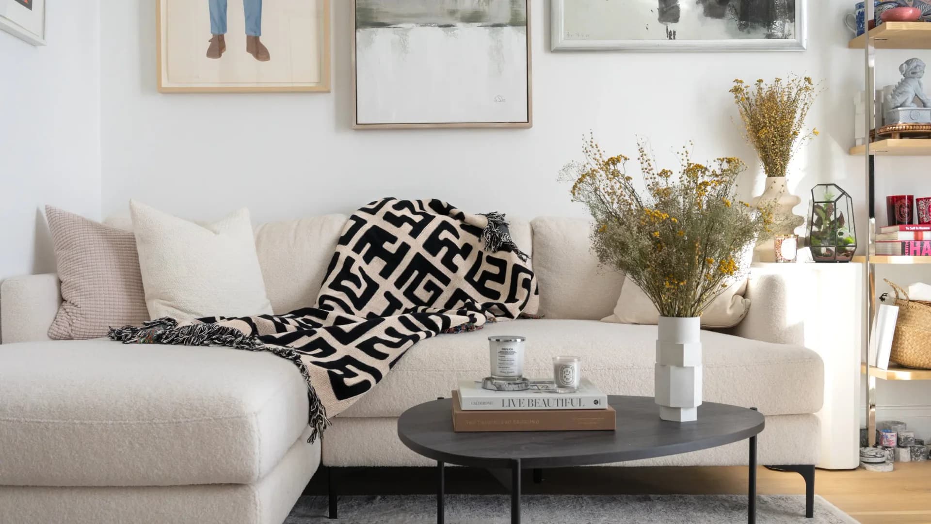 Bright living room with cream sectional sofa, black geometric throw blanket, round black coffee table, and styled shelving with dried florals and decor by Sebastian Bidegain.