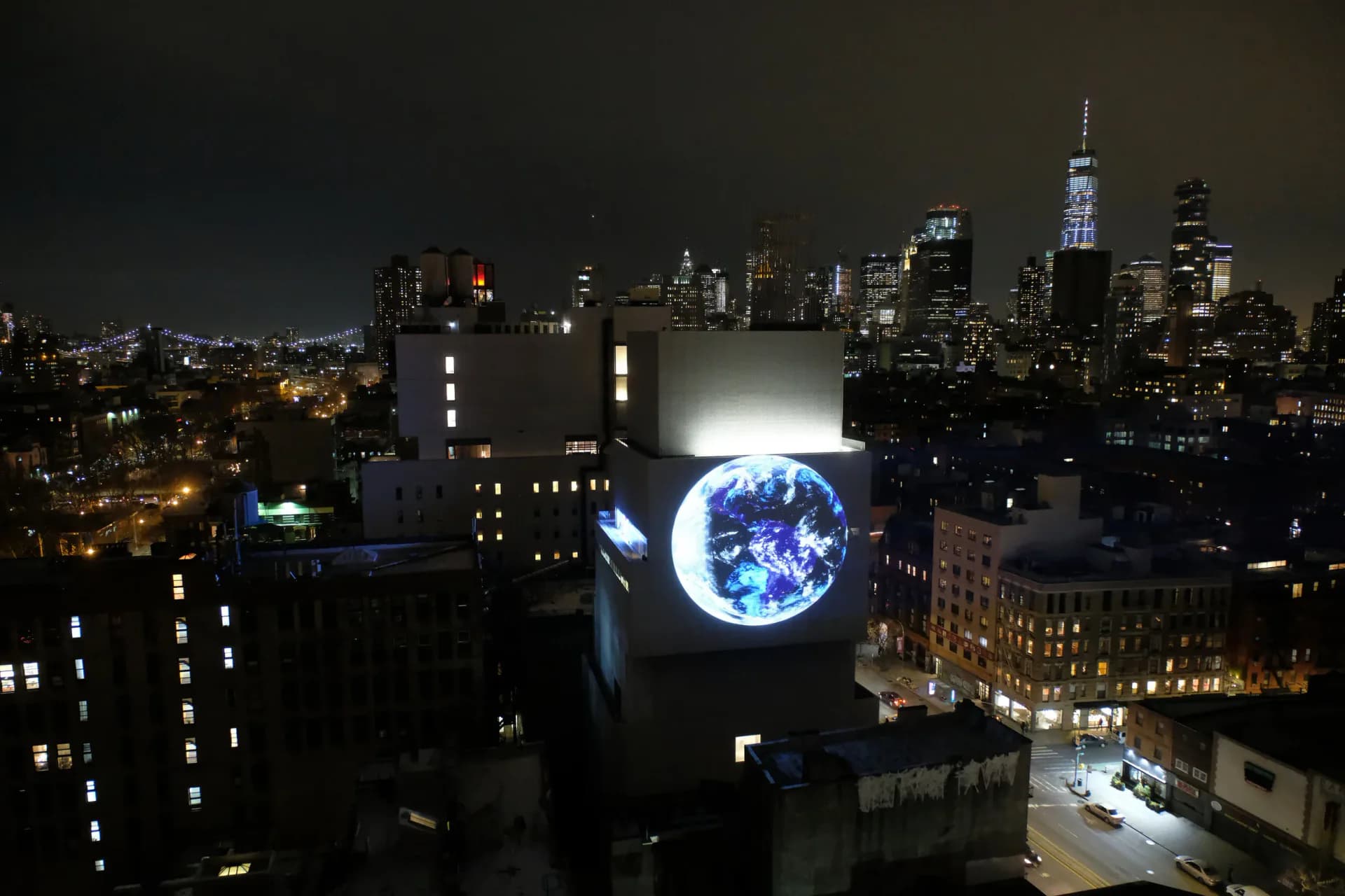 Nighttime NYC skyline view featuring a large illuminated Earth installation suspended between buildings by Sebastian Errazuriz.