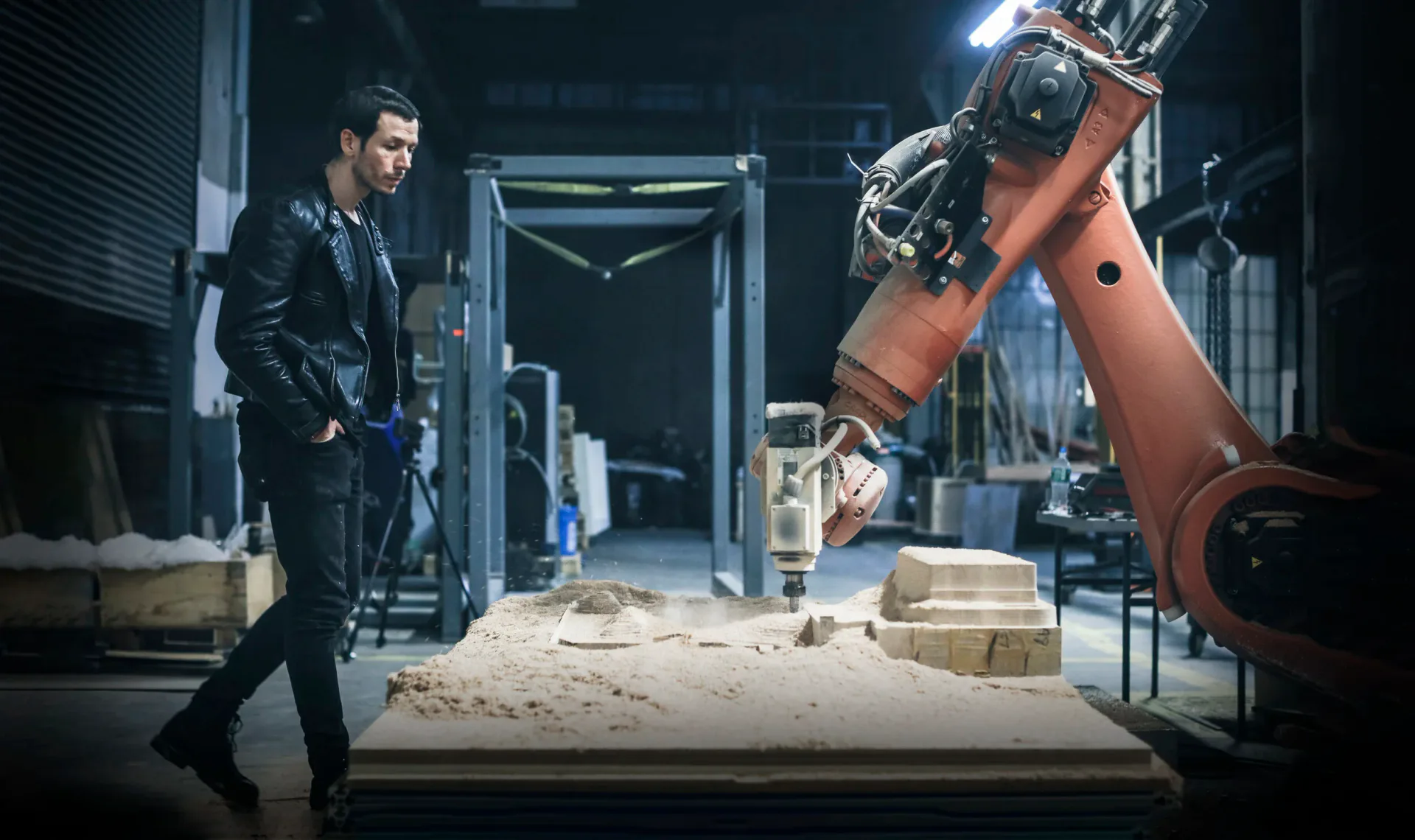 A futuristic workshop interior featuring a man in black leather jacket beside a large robotic arm positioned over a sand-covered table, with industrial lighting and concrete walls by Sebastian Errazuriz.