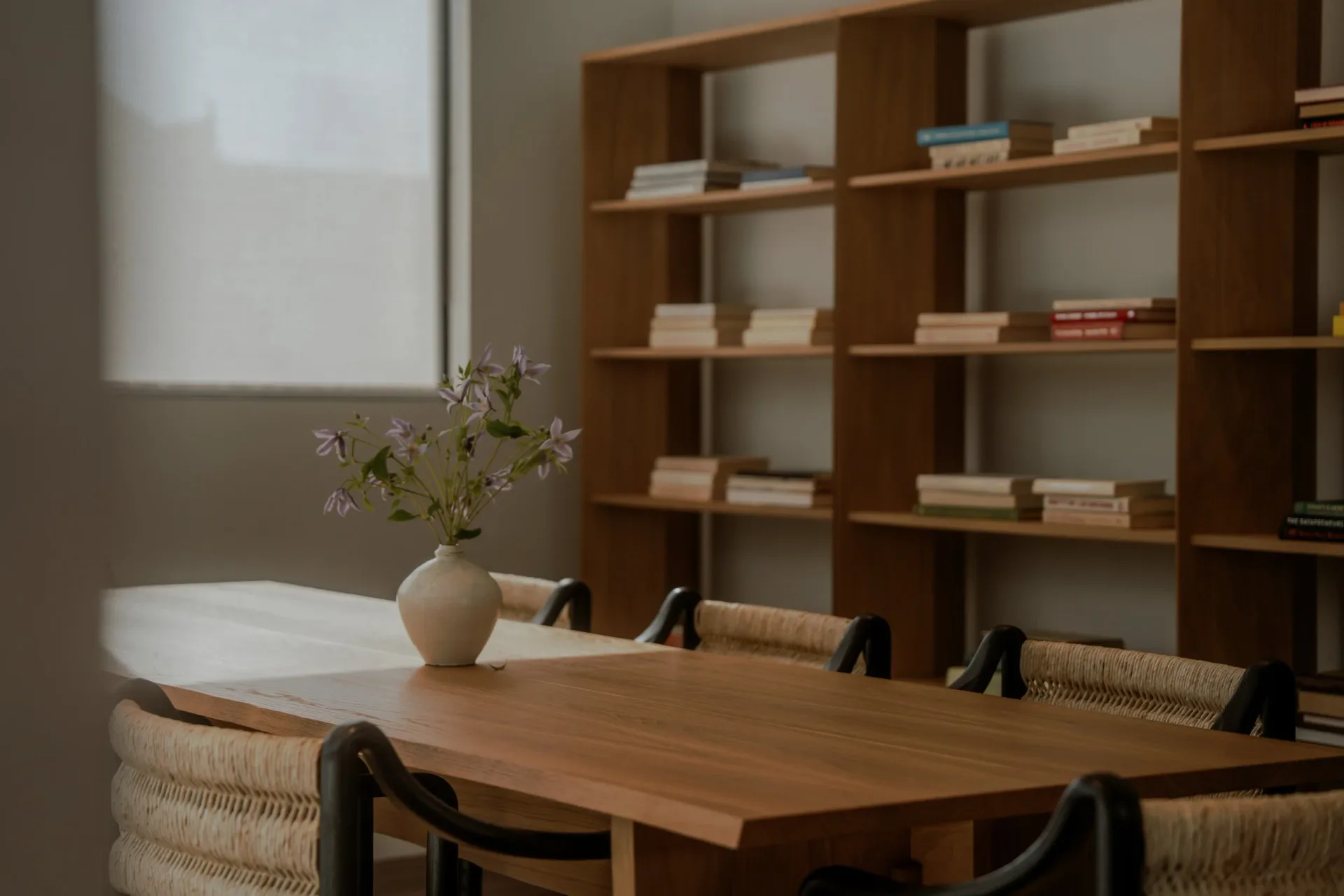 Warm wooden dining table with woven chairs in a minimalist interior featuring built-in shelving and natural light by Sey Jeong.