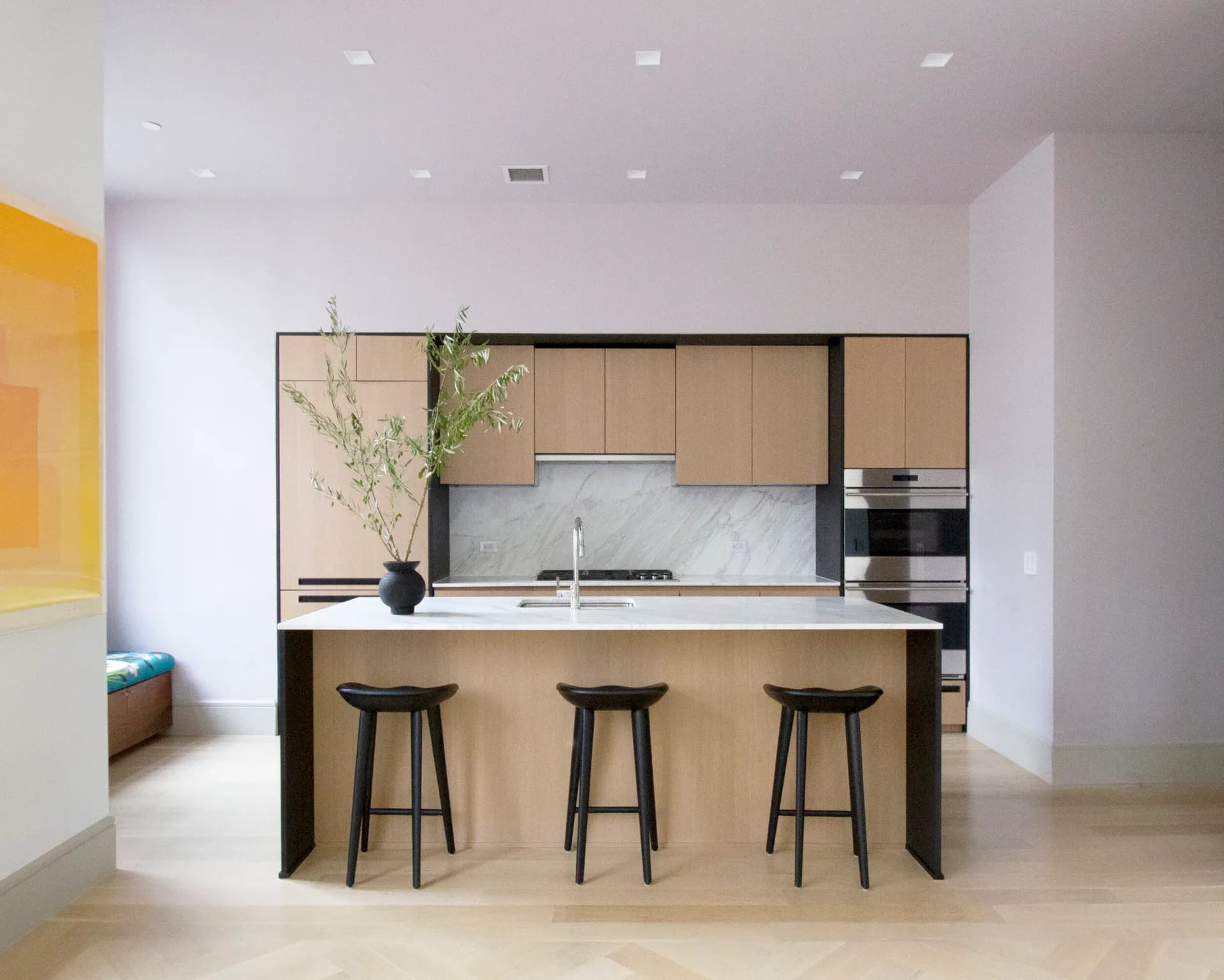 Modern kitchen with light wood cabinetry, marble backsplash, black bar stools, and minimalist fixtures designed by Sey Jeong.