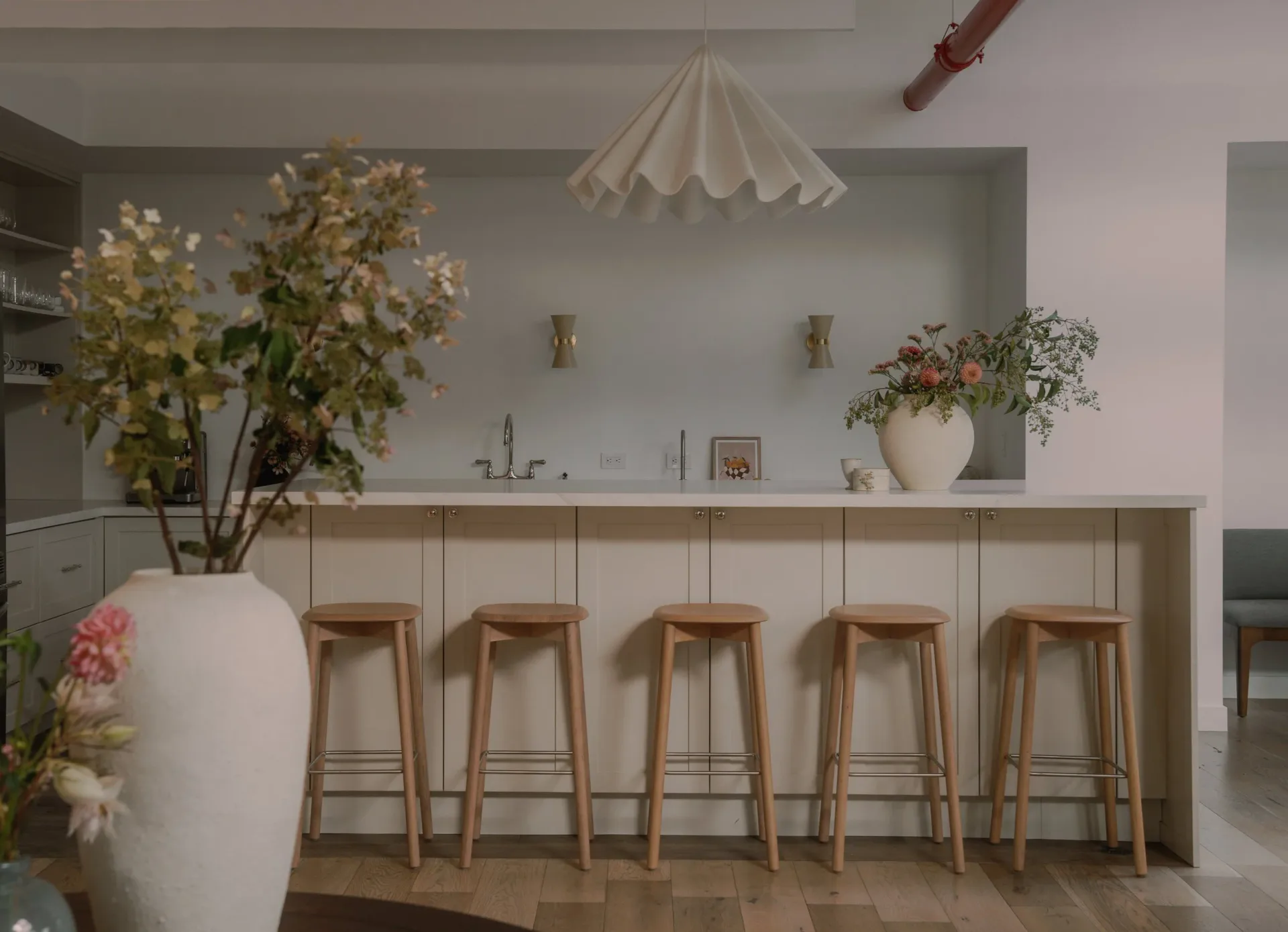Minimalist kitchen bar with five wooden stools and white countertop, fresh flower arrangements, and soft neutral palette by Sey Jeong.