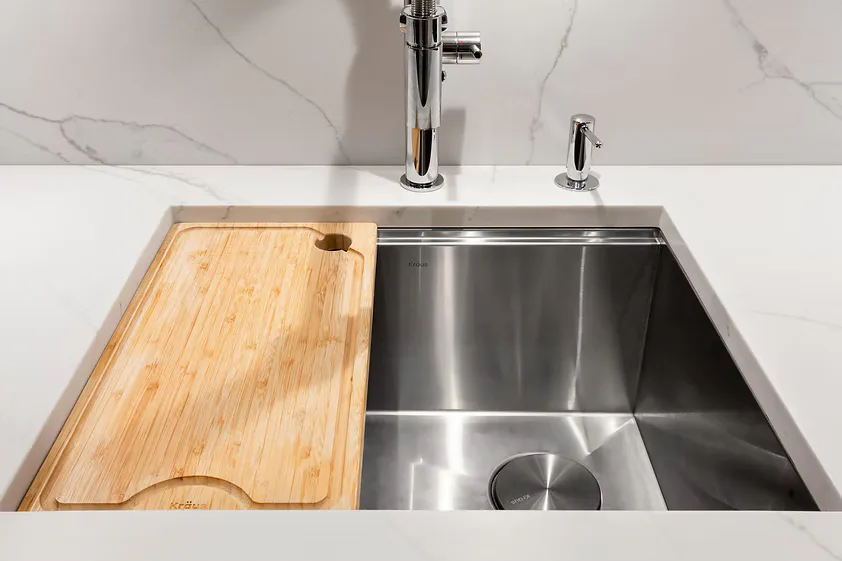 Modern kitchen sink with stainless steel double basin, wooden cutting board, and marble backsplash designed by Shai Arviv.