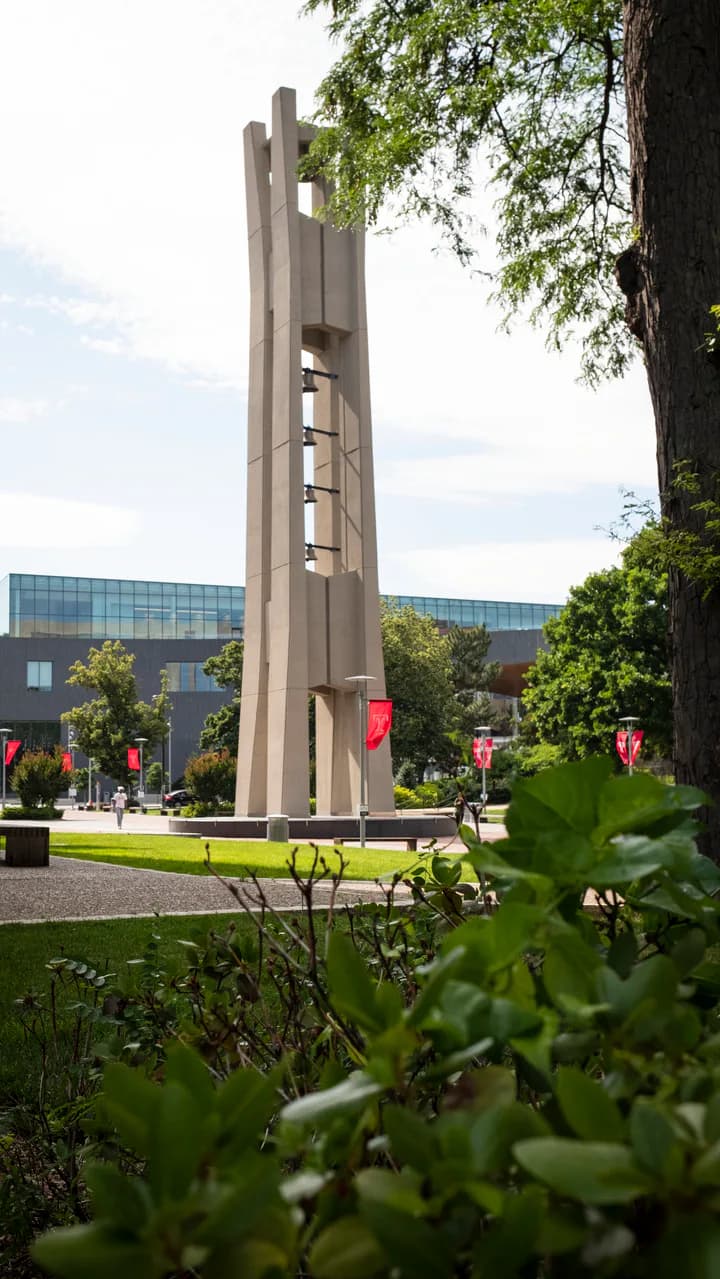 Tall concrete modernist tower with vertical window openings in a landscaped plaza with red banners, designed by Shalin Dave.