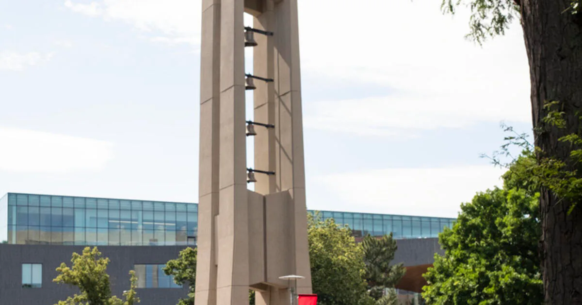 Modernist concrete bell tower with horizontal banding stands beside contemporary glass-facade building surrounded by mature trees, by Shalin Dave.