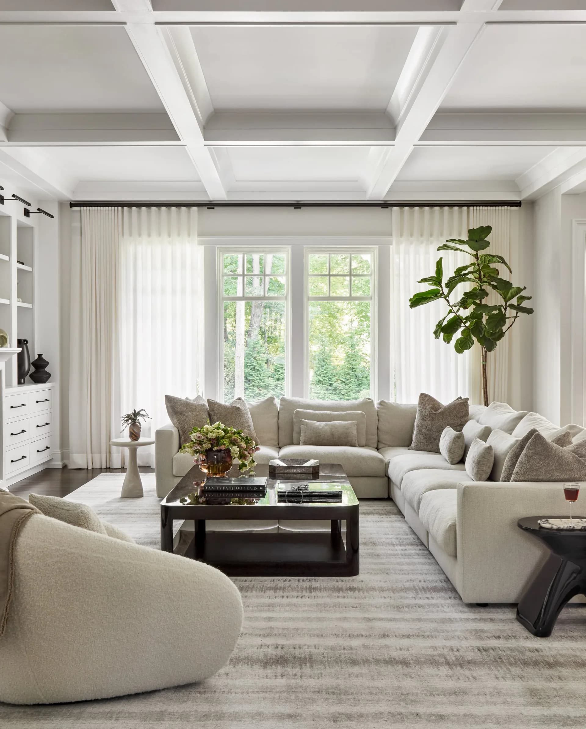 Elegant living room with coffered ceiling, neutral linen seating, dark wood coffee table, and large windows overlooking greenery by Shana Shaiman.