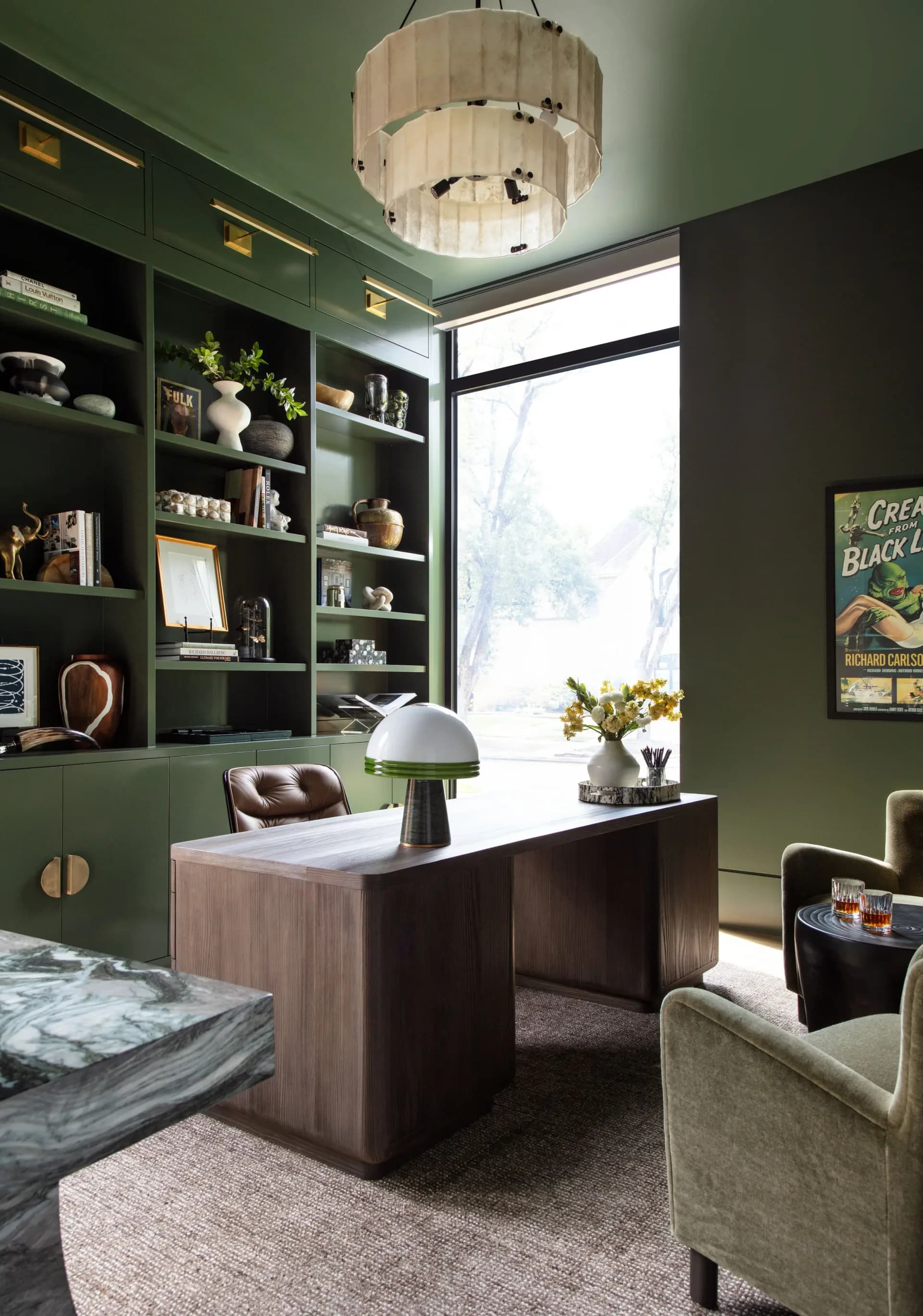Modern home office with forest-green built-in shelving, wooden desk, pendant light fixture, and large windows overlooking nature by Shana Shaiman.