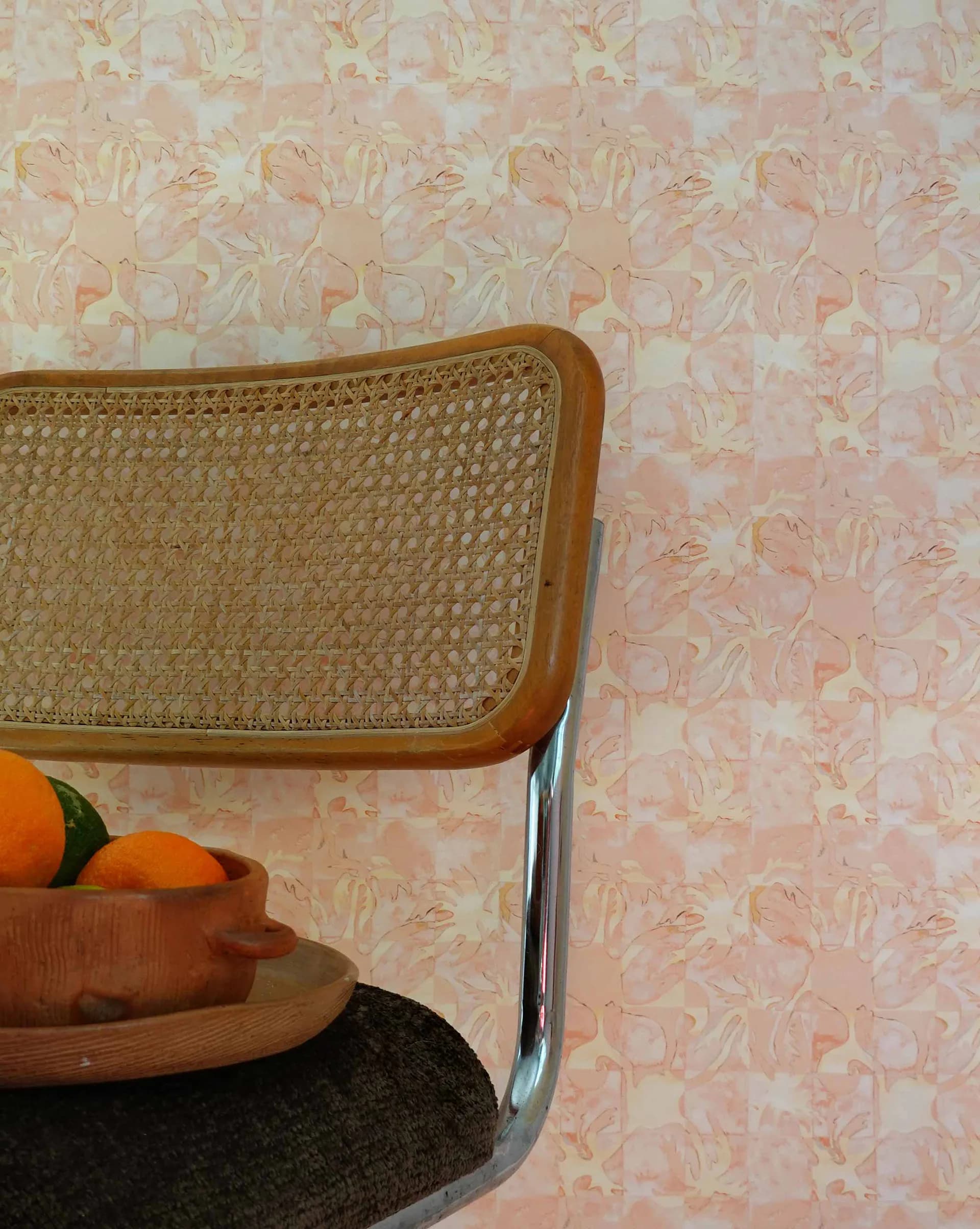 Mid-century modern interior detail featuring a caned chair with chrome base against pink floral wallpaper, styled with wooden bowl and citrus fruit by Shanan Campanaro.