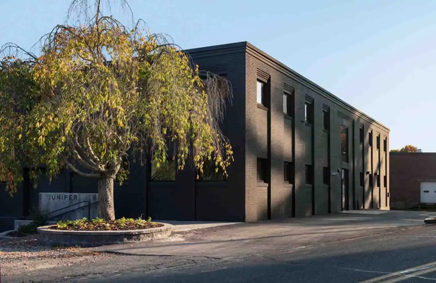 Contemporary brick industrial building with dark facade, large windows, and weeping willow tree, photographed in natural daylight by Shant Madjarian.