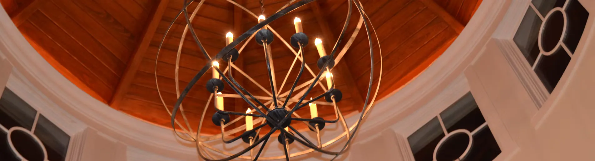 Modern ceiling installation featuring a brass orb chandelier with geometric wireframe design against warm wood soffit and neutral walls by Sharon Nizolek.
