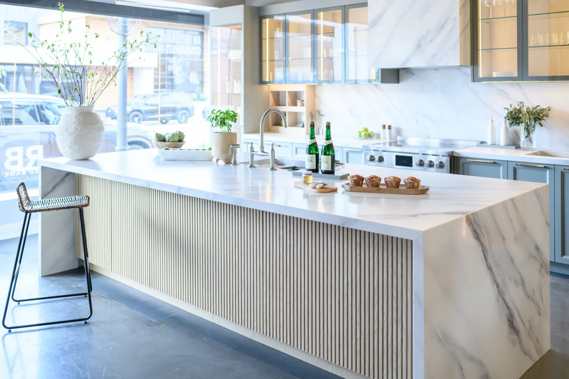 Modern kitchen with white marble island featuring vertical ribbed front panel, white subway backsplash, blue cabinetry, and large windows overlooking city by Shayna Vrabel.