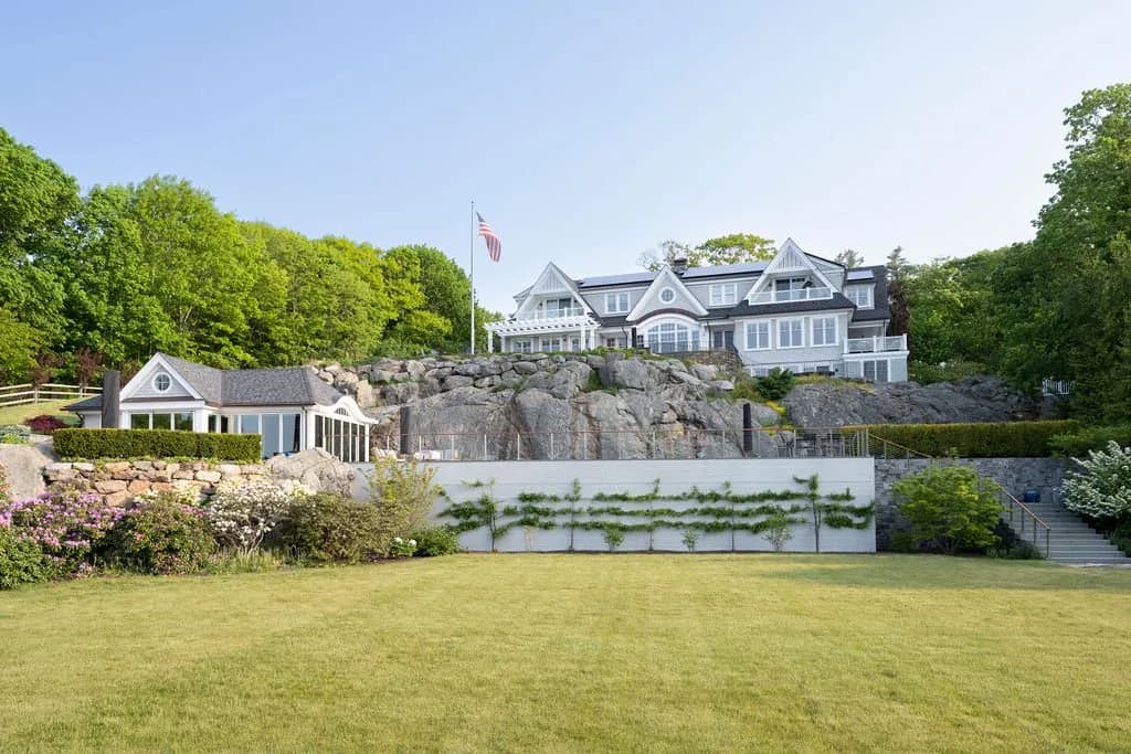 Waterfront residential property featuring a large multi-story home perched on rocky outcropping with manicured lawn, stone walls, and modern architectural details by Shayna Vrabel.